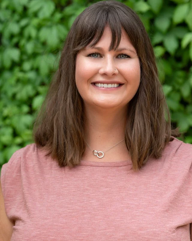 A smiling woman with shoulder-length brown hair and blue eyes, wearing a pink top and a silver necklace, standing outdoors with green leaves in the background in Texas