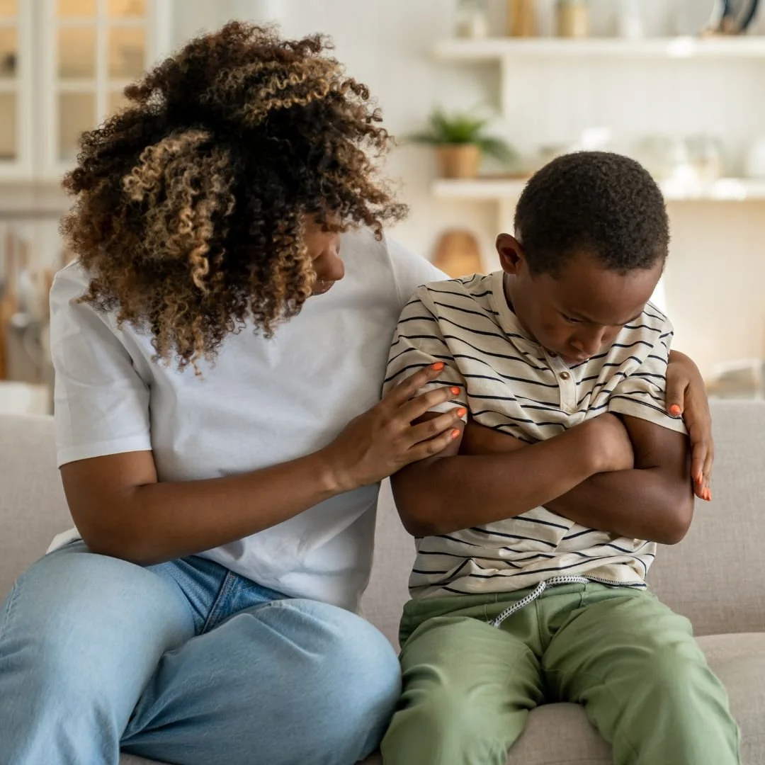 A woman comforts a teenage boy who has crossed his arms and appears upset, sitting together on a sofa in a cozy living room.
