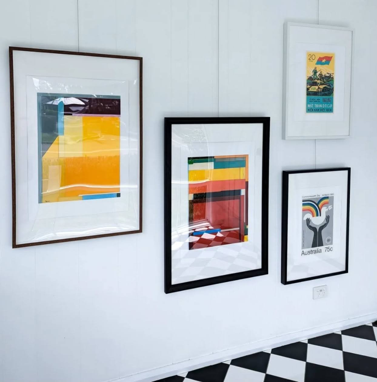 Series of framed artworks on a white wall, including abstract colorful patterns and a postage stamp with an illustration of a rainbow and hands, against a black and white diamond-patterned floor.