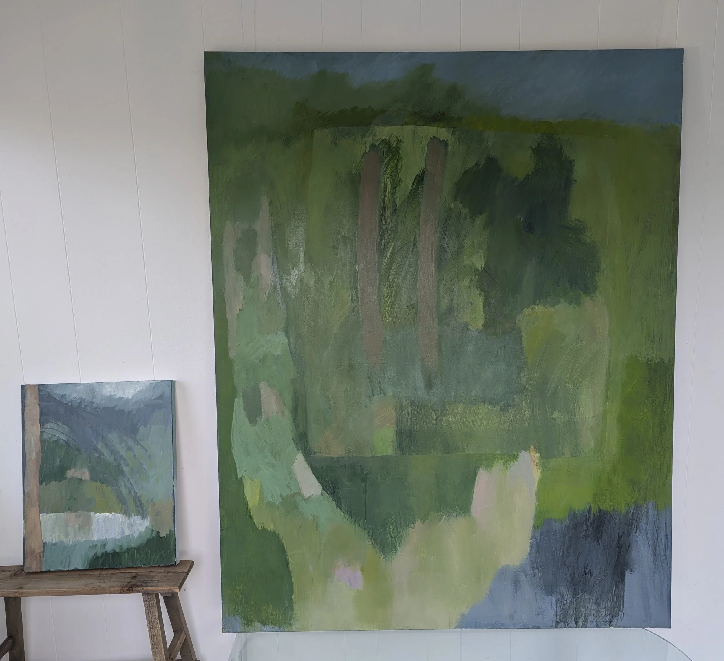 Two abstract landscape paintings in shades of green, blue, and beige; one large painting leaning against a white wall, and a smaller one on a wooden stool.