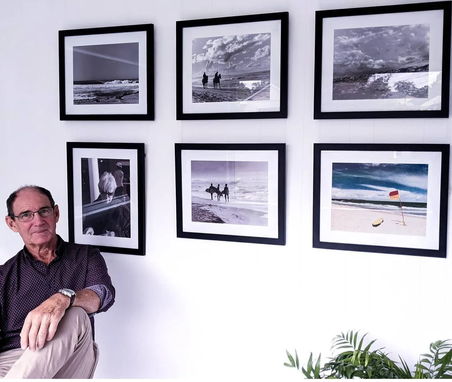 A man sitting in front of a wall with six framed photographs of beach scenes, some in black and white and some in color, with greenery at the bottom right.
