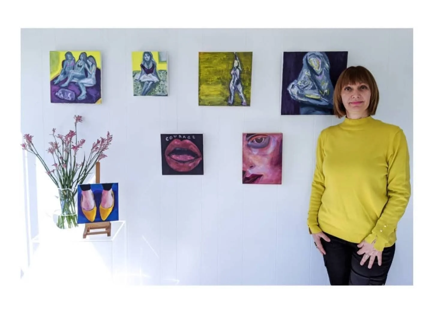A woman with short brown hair wearing a bright yellow turtleneck sweater and black pants standing in front of a white wall with various colorful paintings. A vase with pink flowers is on a small table next to her.