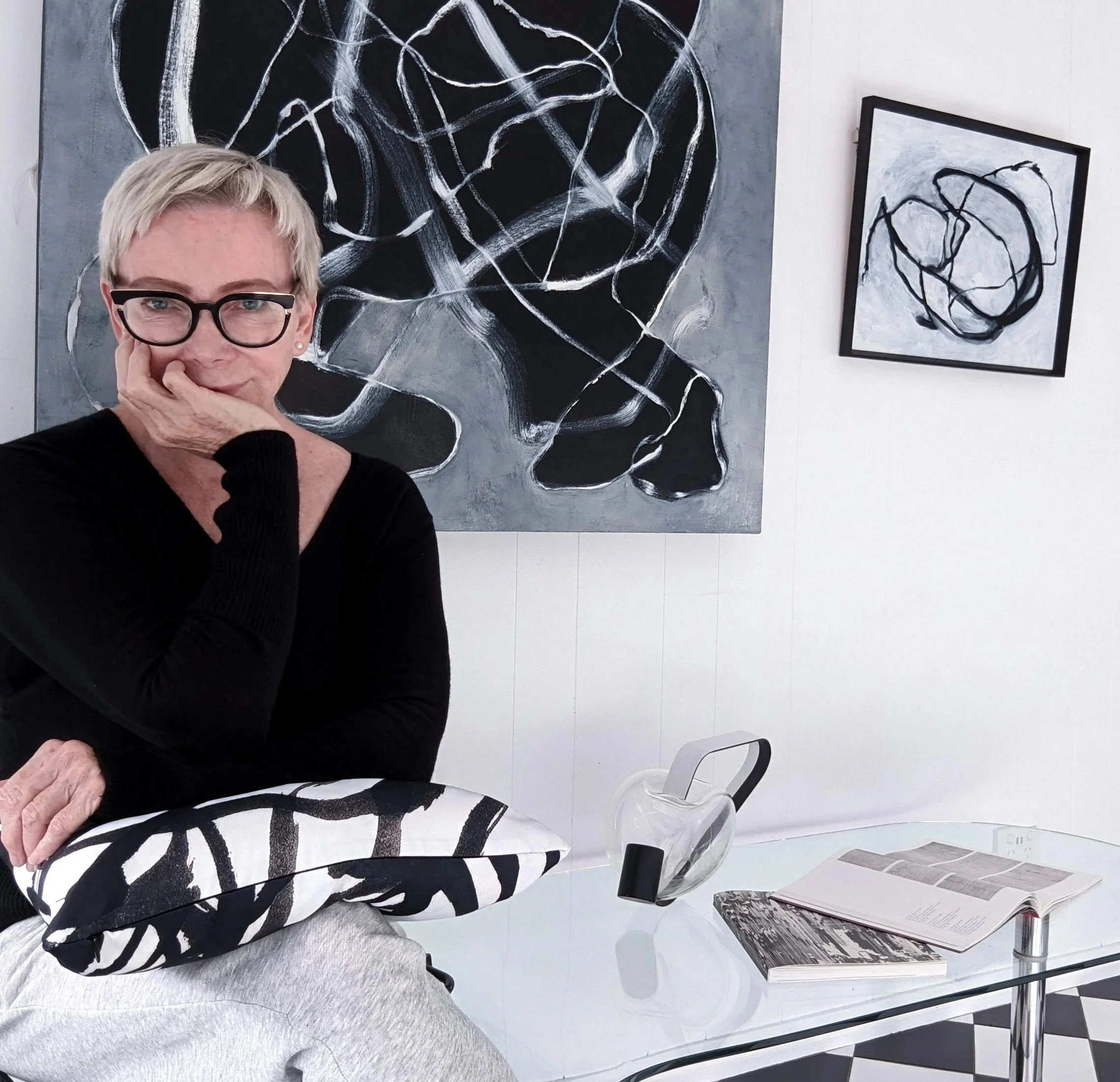 A woman with short blonde hair, wearing glasses and a black top, sitting in an art gallery. She is resting her chin on her hand, with a pillow featuring black and white abstract patterns on her lap. Behind her are two abstract black and white paintings on the wall.