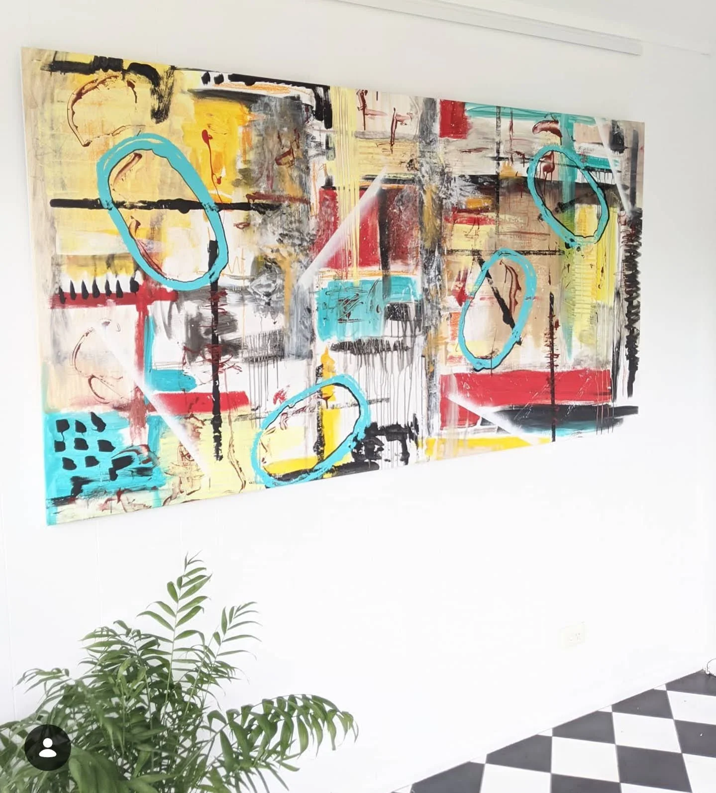 Abstract colorful painting on a white wall with blue circles and dynamic lines, with a green plant in the foreground and black and white checkered flooring.