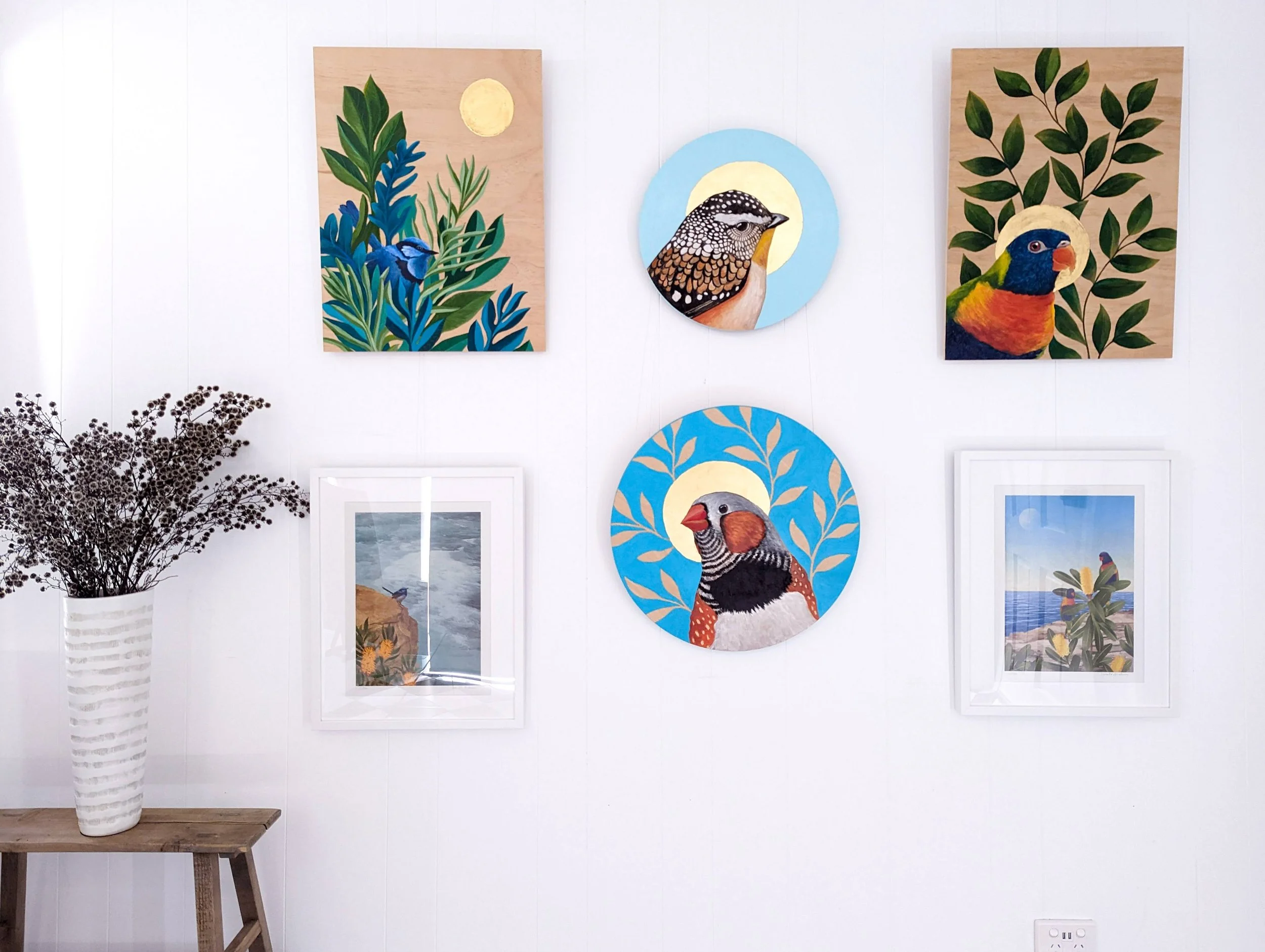 A white wall decorated with colorful bird-themed artwork and a framed photograph. To the left, there is a wooden table with a white vase holding dried branches.