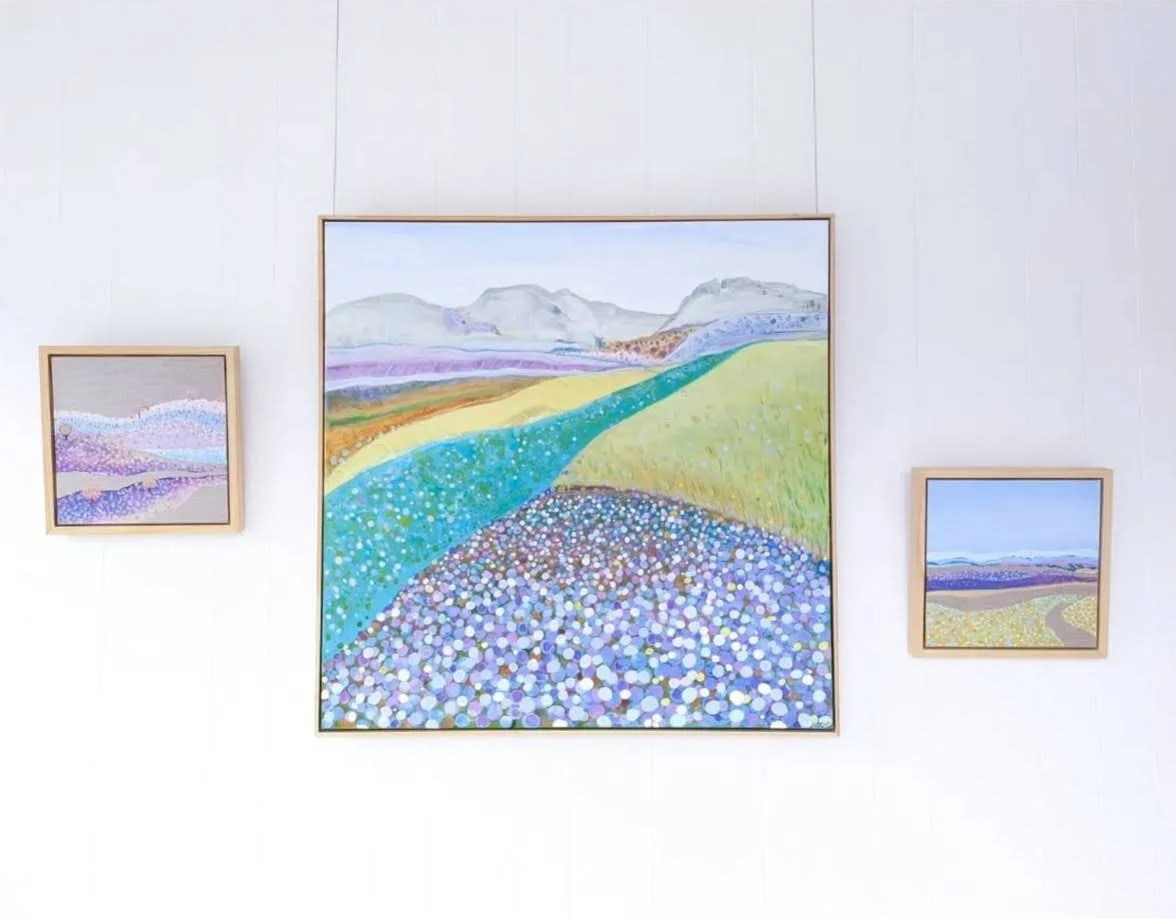 Three landscape paintings on a white wall, with the largest in the center depicting colorful rolling hills and fields in an abstract style.