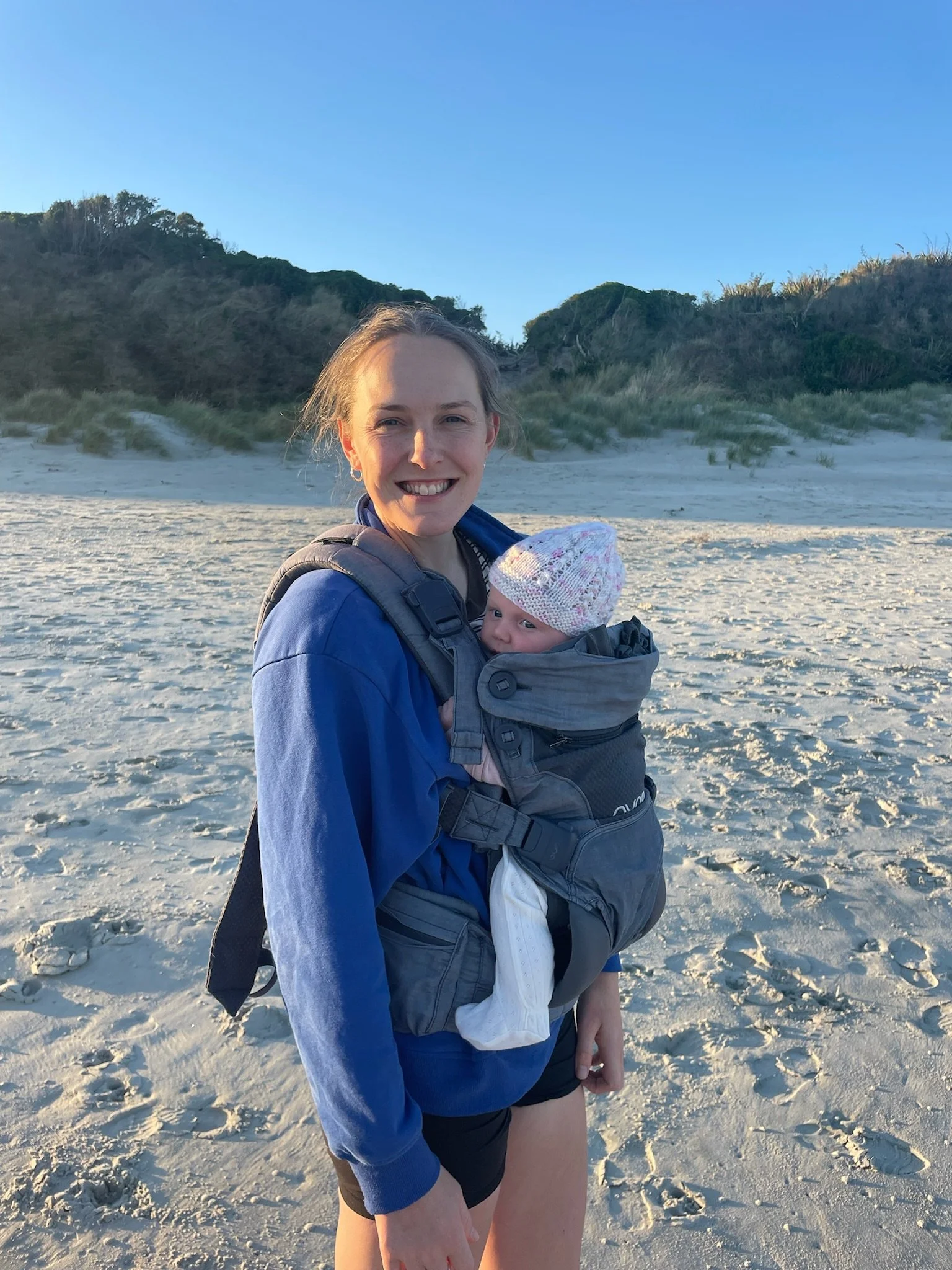A woman smiling while holding a baby in a carrier on a sandy beach with dunes and green vegetation in the background.