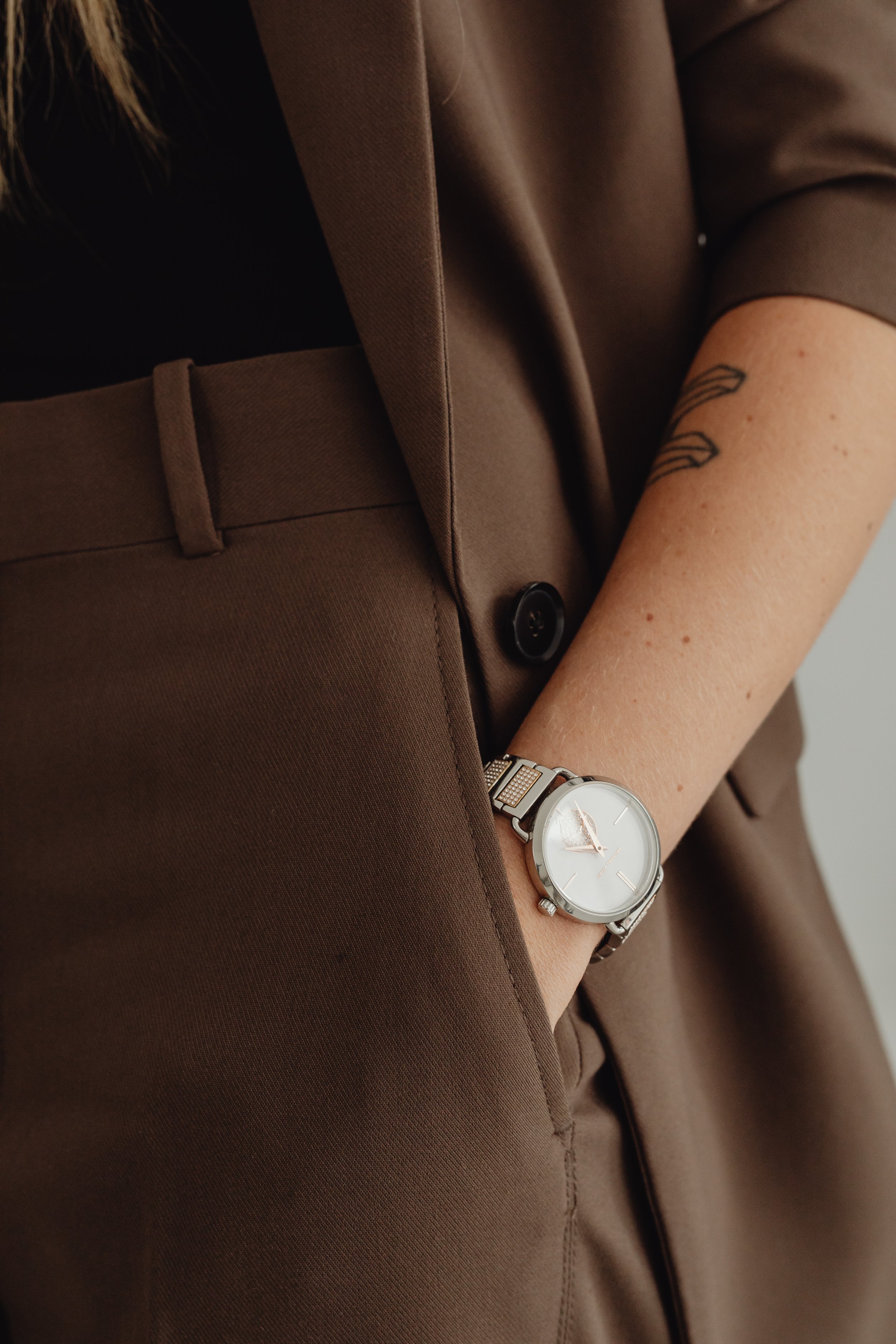 Fashionable woman in a chocolate brown suit, closely cropped to show her hand in her pocket with a watch and arm tattoo