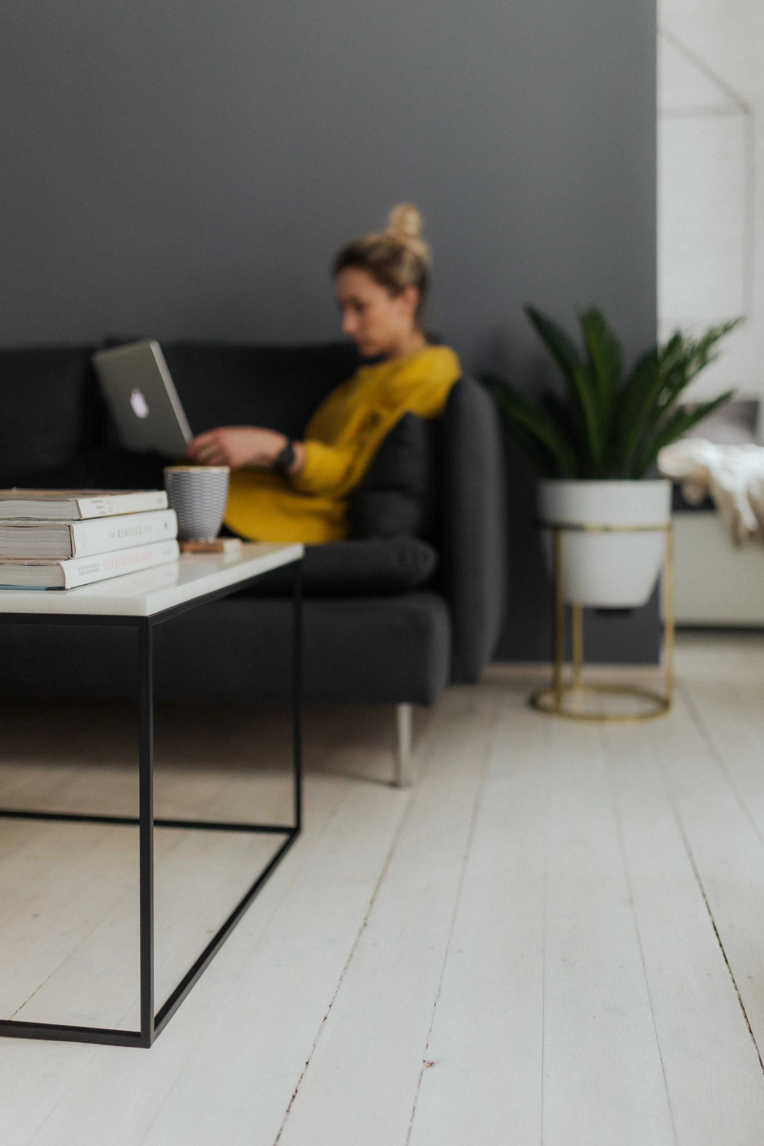 Coffee table with book in the foreground with a woman sitting on her couch working on her laptop in the background