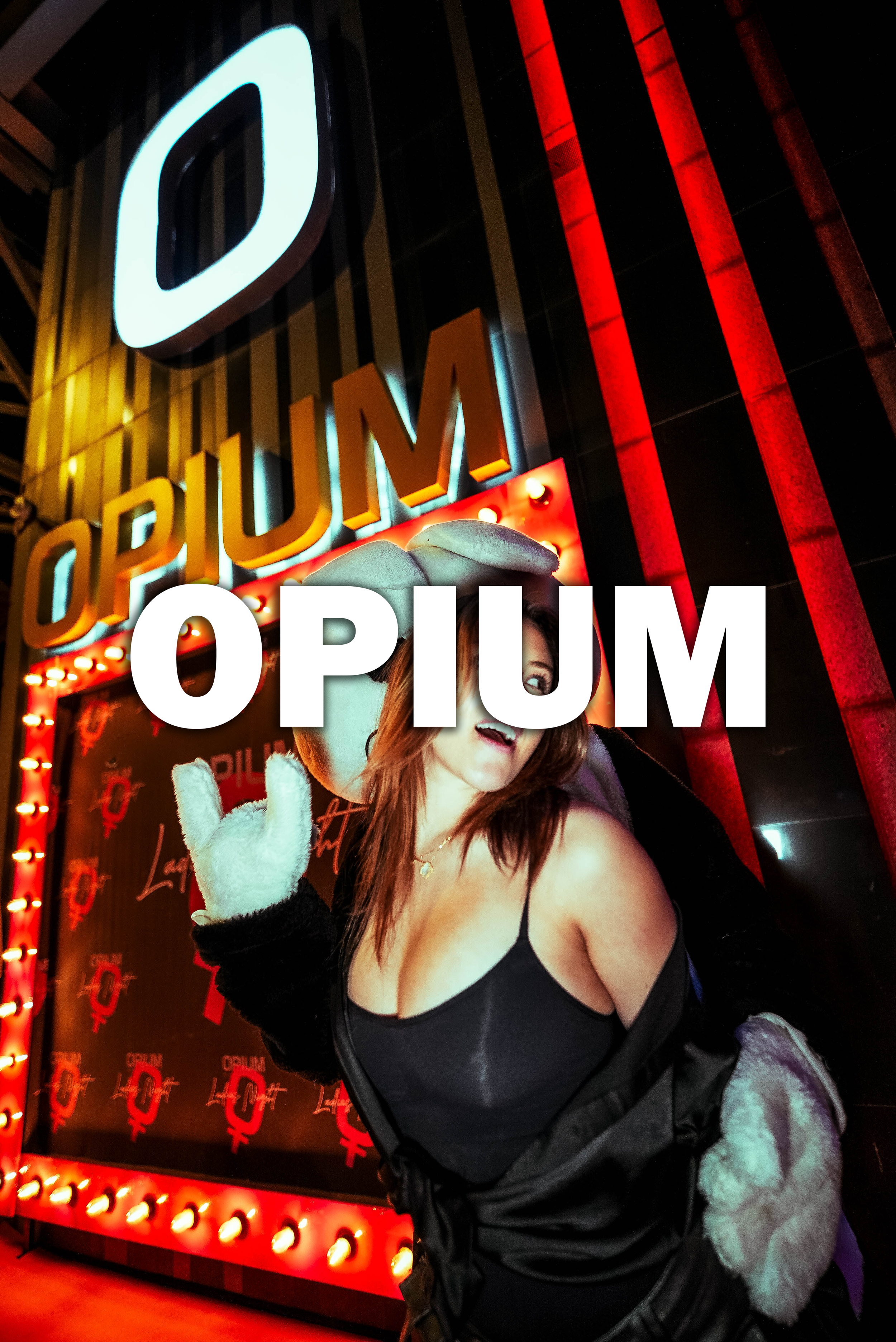 A woman wearing a black dress and a white furry coat posing in front of a neon-lit sign that reads 'OPIUM'. She has a playful expression, is making a peace sign with her hand, and is standing near the entrance of a club or entertainment venue with red lighting and decorative elements.