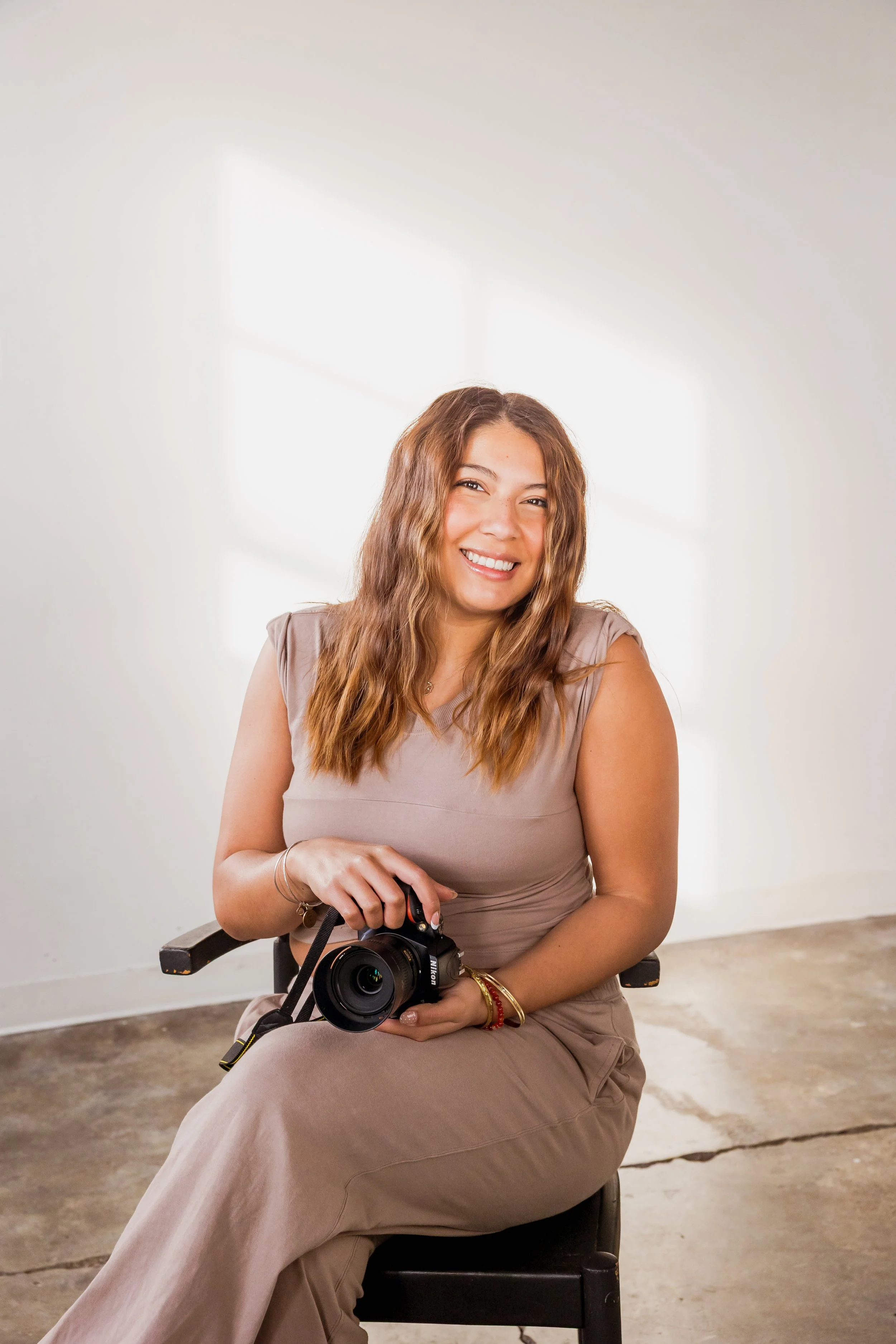 Portrait of a smiling woman sitting on a black chair, holding a camera, with a shadow of window panes cast on the white wall behind her.