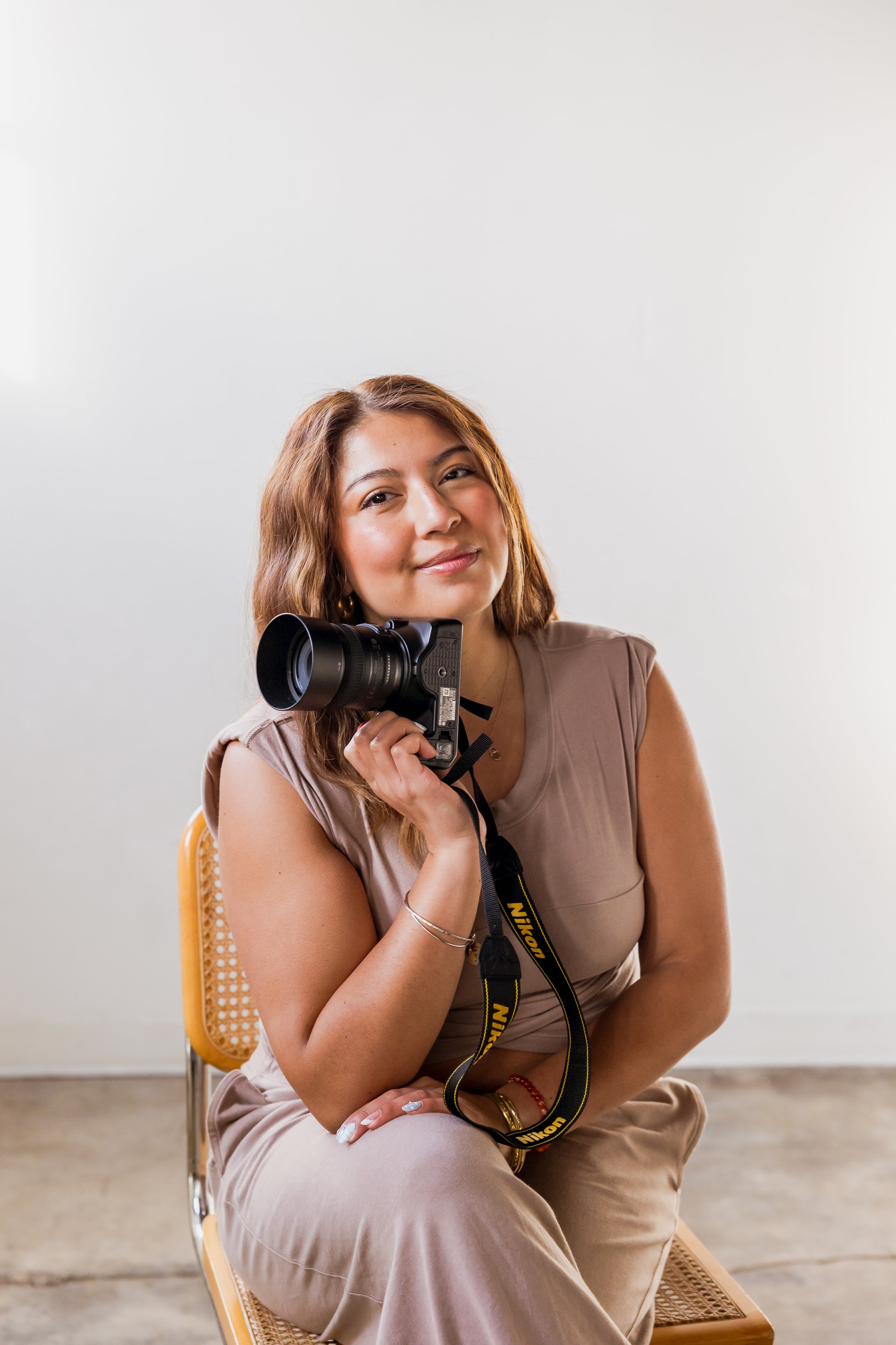 A woman sitting on a chair holding a Nikon camera, smiling, in front of a plain white wall.