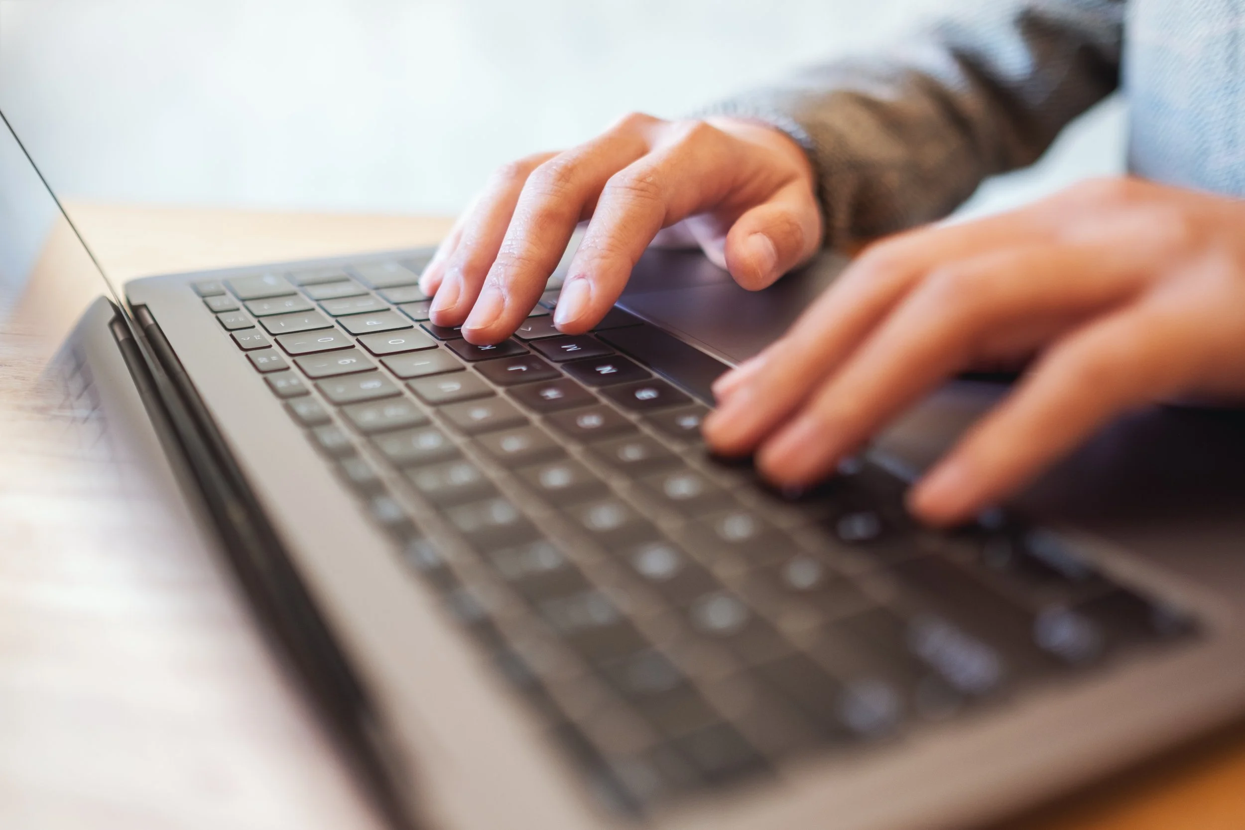 Close-up of a person typing on a laptop keyboard with their right hand, wearing a long sleeve shirt, on a wooden surface.