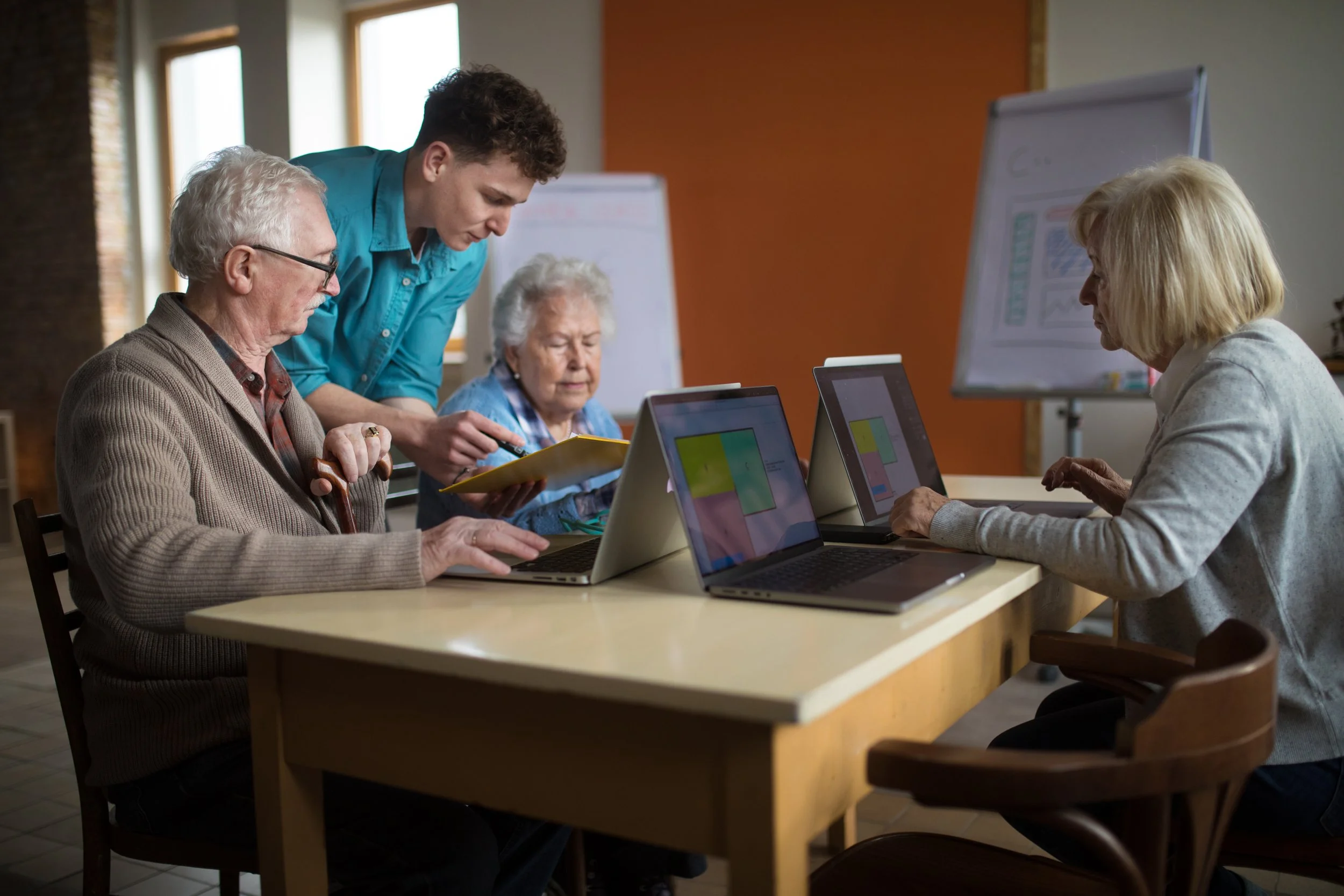 A group of five people, including elderly men and women and a younger man, gathered around a wooden table working on laptops, with a woman in the background looking at a tablet. The room has large windows and a whiteboard.