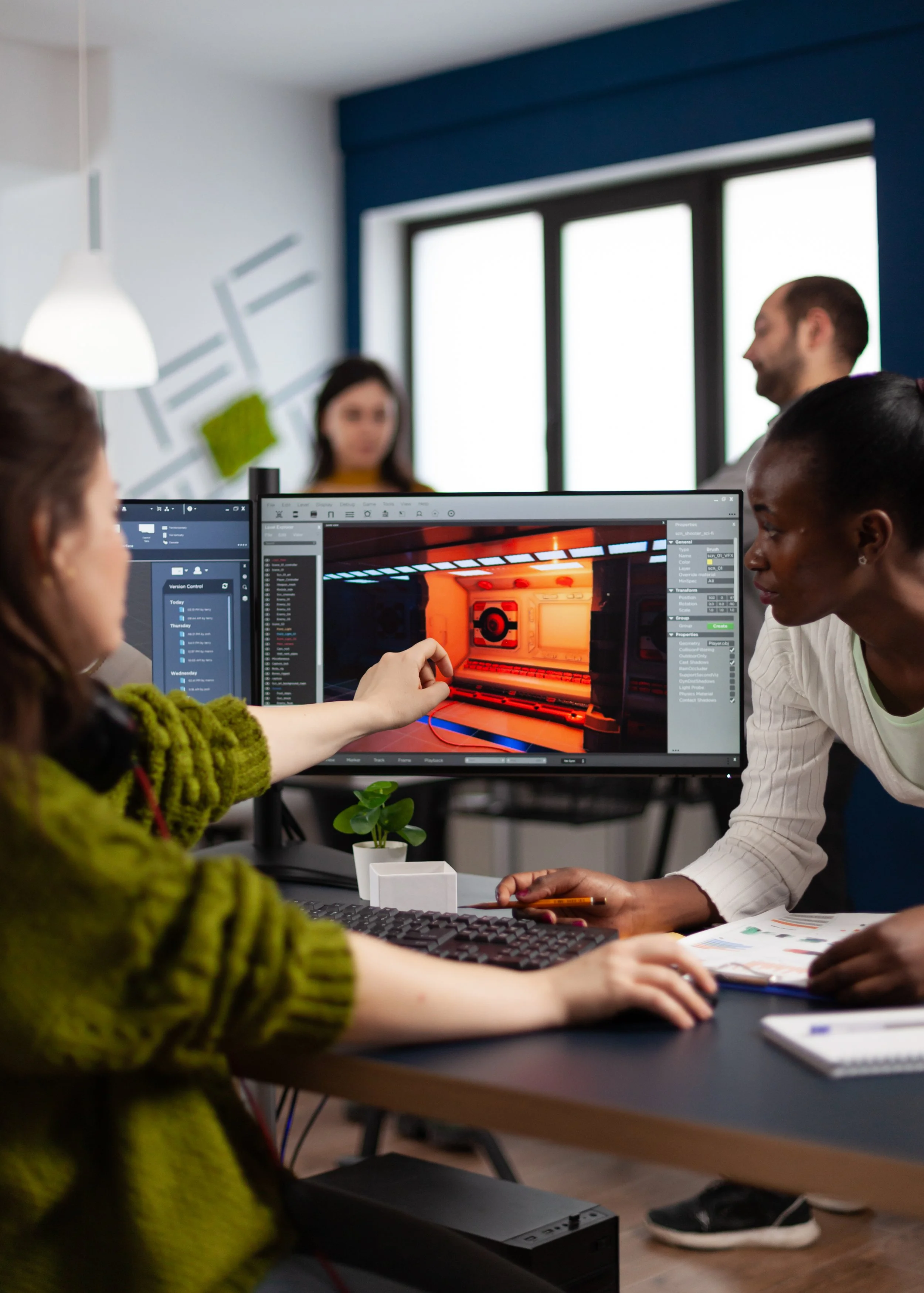 Four people working together in a modern office, two women and one man sitting at a desk with computer screens, collaborating on a 3D design project displayed on the monitor. In the background, another woman is standing near a wall with geometric wall art, with a large window providing natural light.