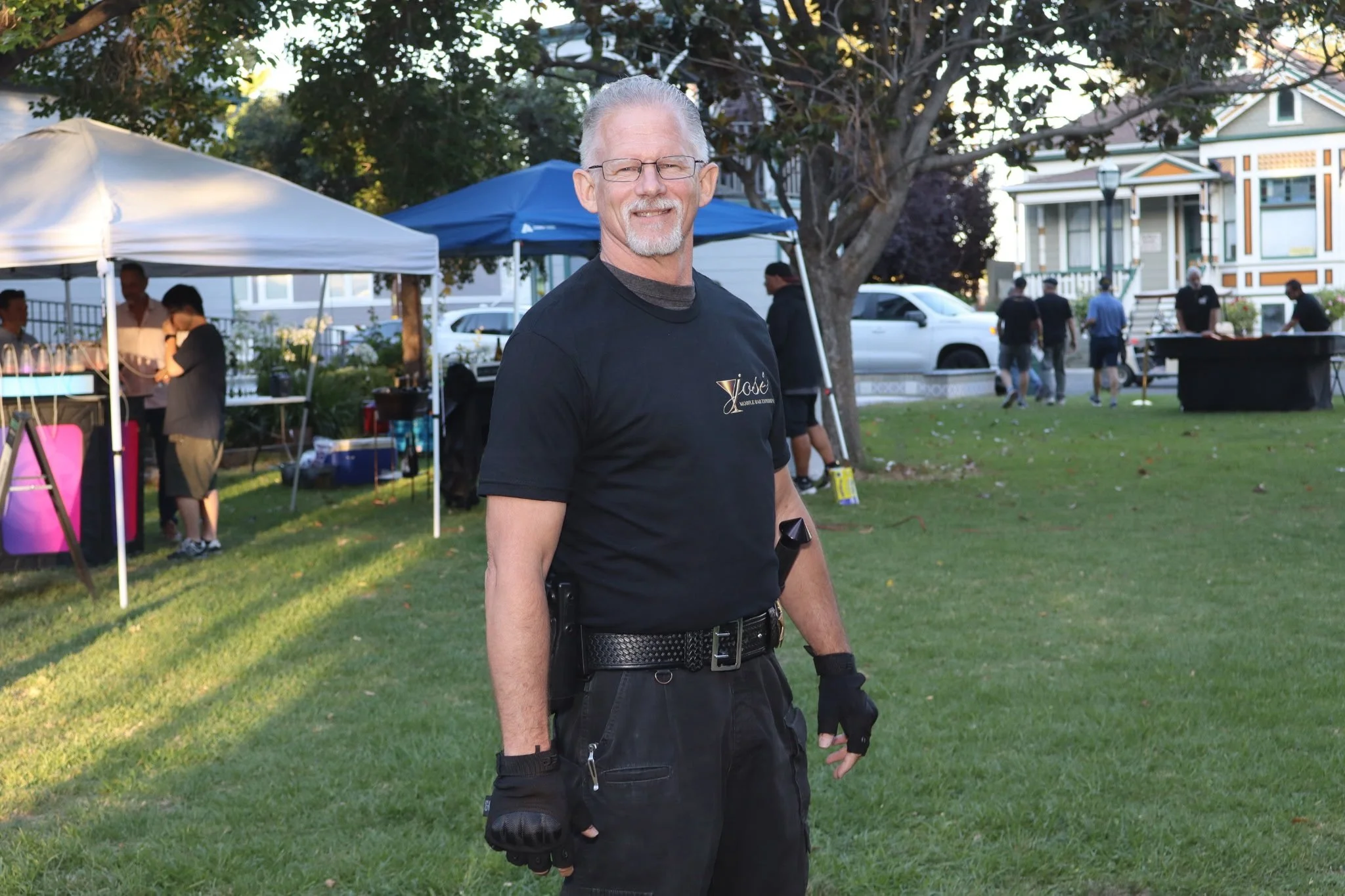A middle-aged man with gray hair, glasses, and a goatee, wearing a black police officer uniform, standing outdoors on a grassy area during a community event. There are tents, people, and houses in the background.