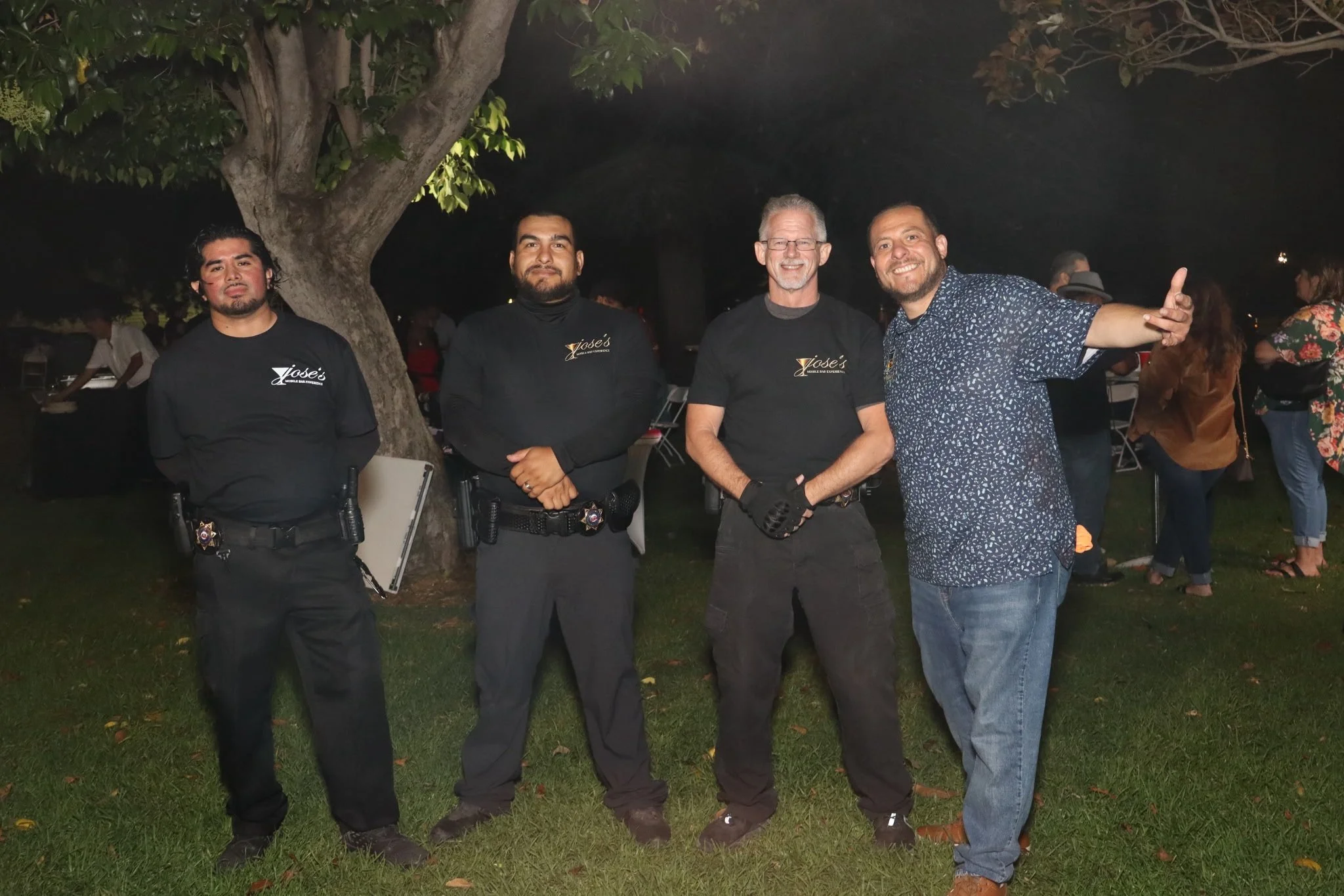 Four men standing outdoors at night, three wearing black shirts with a logo that says 