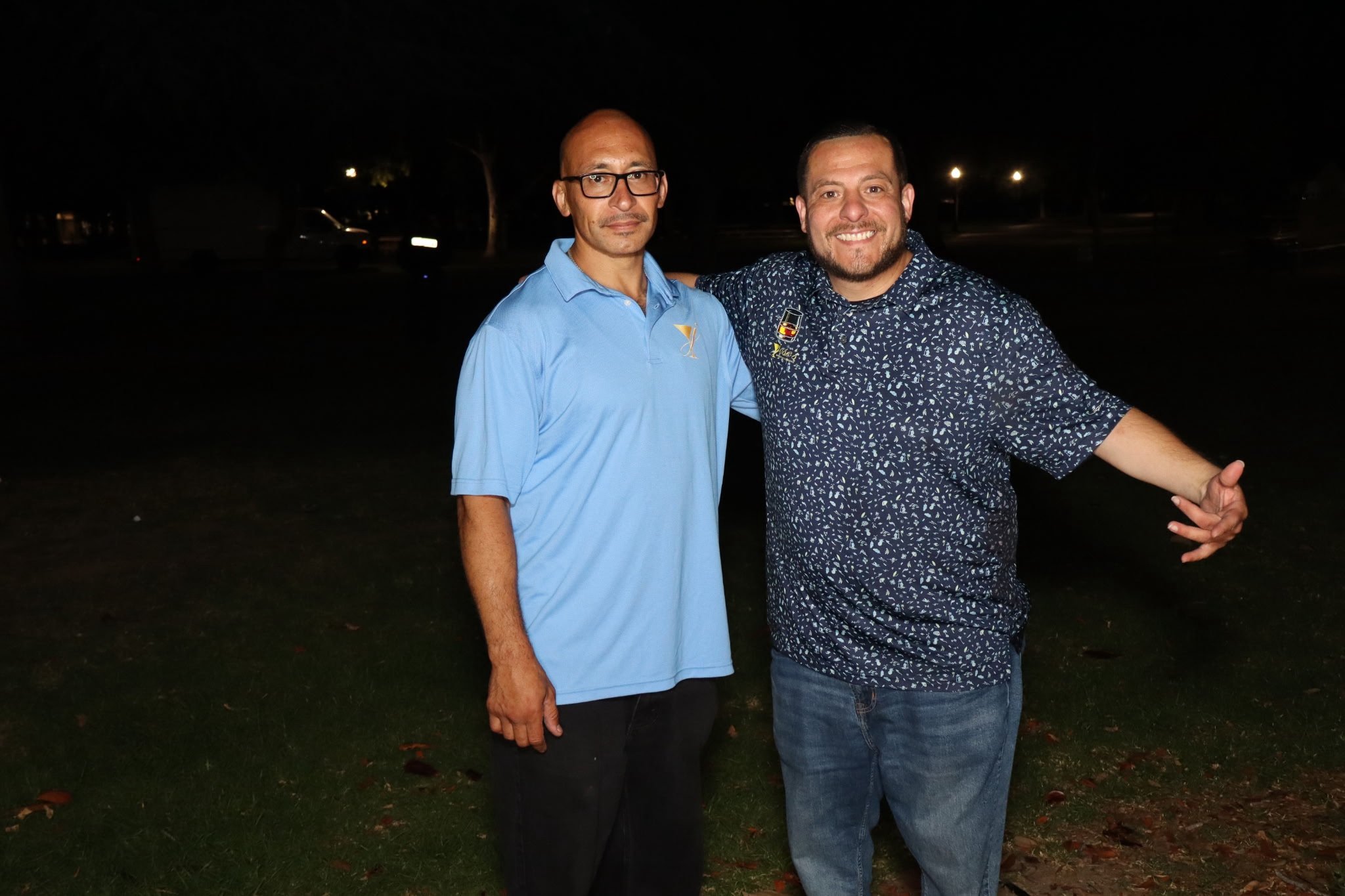 Two men standing outdoors at night, one with glasses and a light blue shirt, the other with a patterned shirt, smiling, with trees and streetlights in the background.