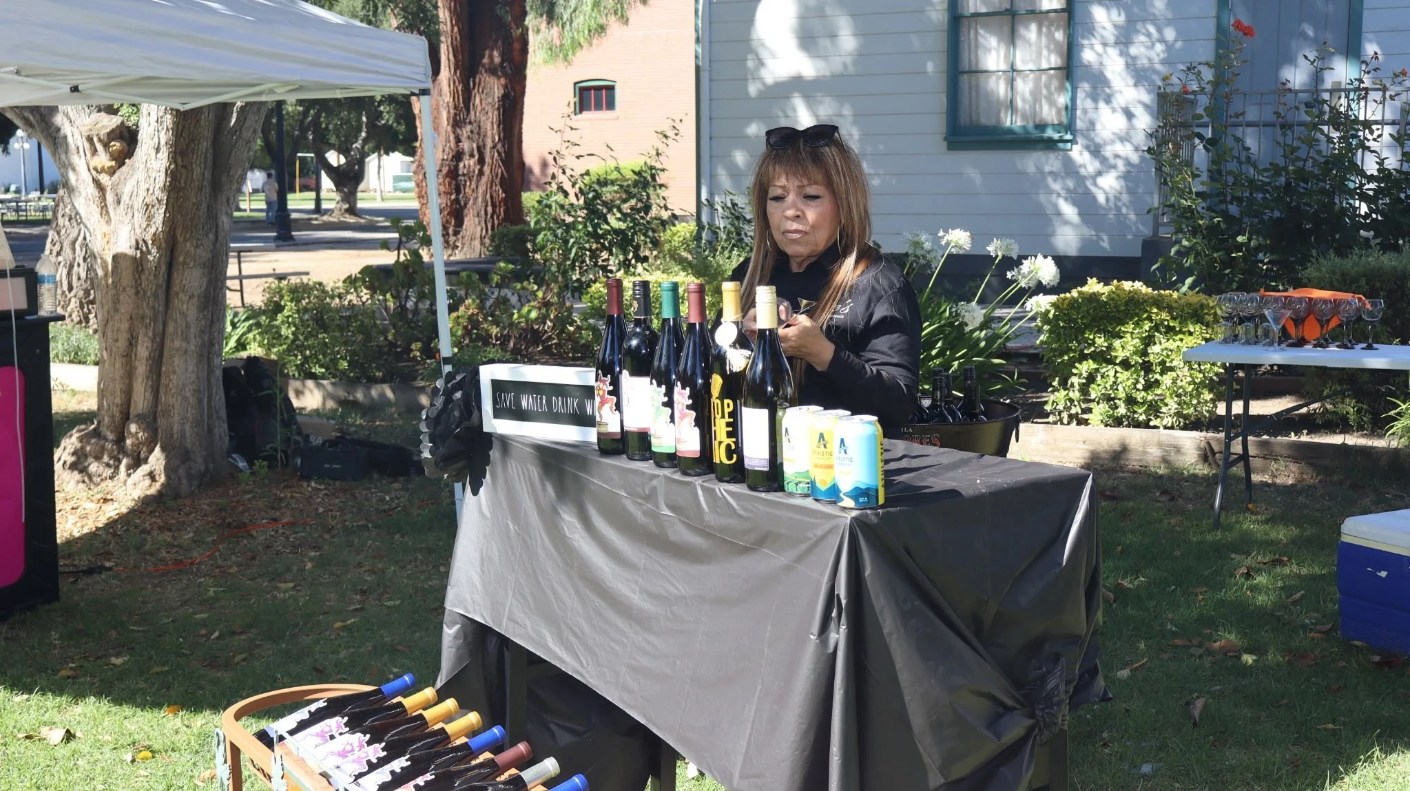A woman with sunglasses on her head standing behind a table with bottles of wine and cans, outdoors in a park with trees, bushes, and a house in the background.