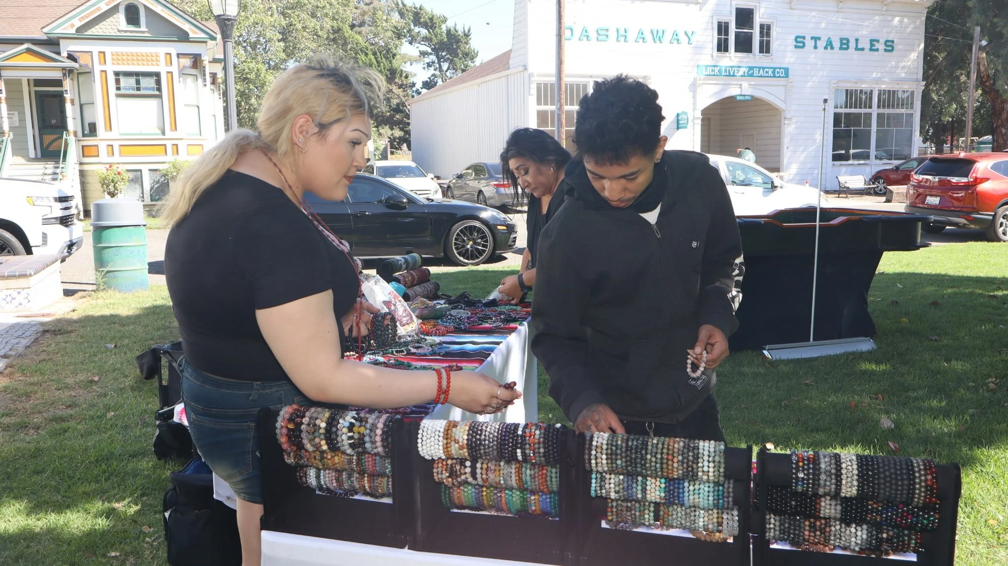 People browsing jewelry and accessories at an outdoor market stall with a white building labeled 'DASH AWAY STABLES' in the background.