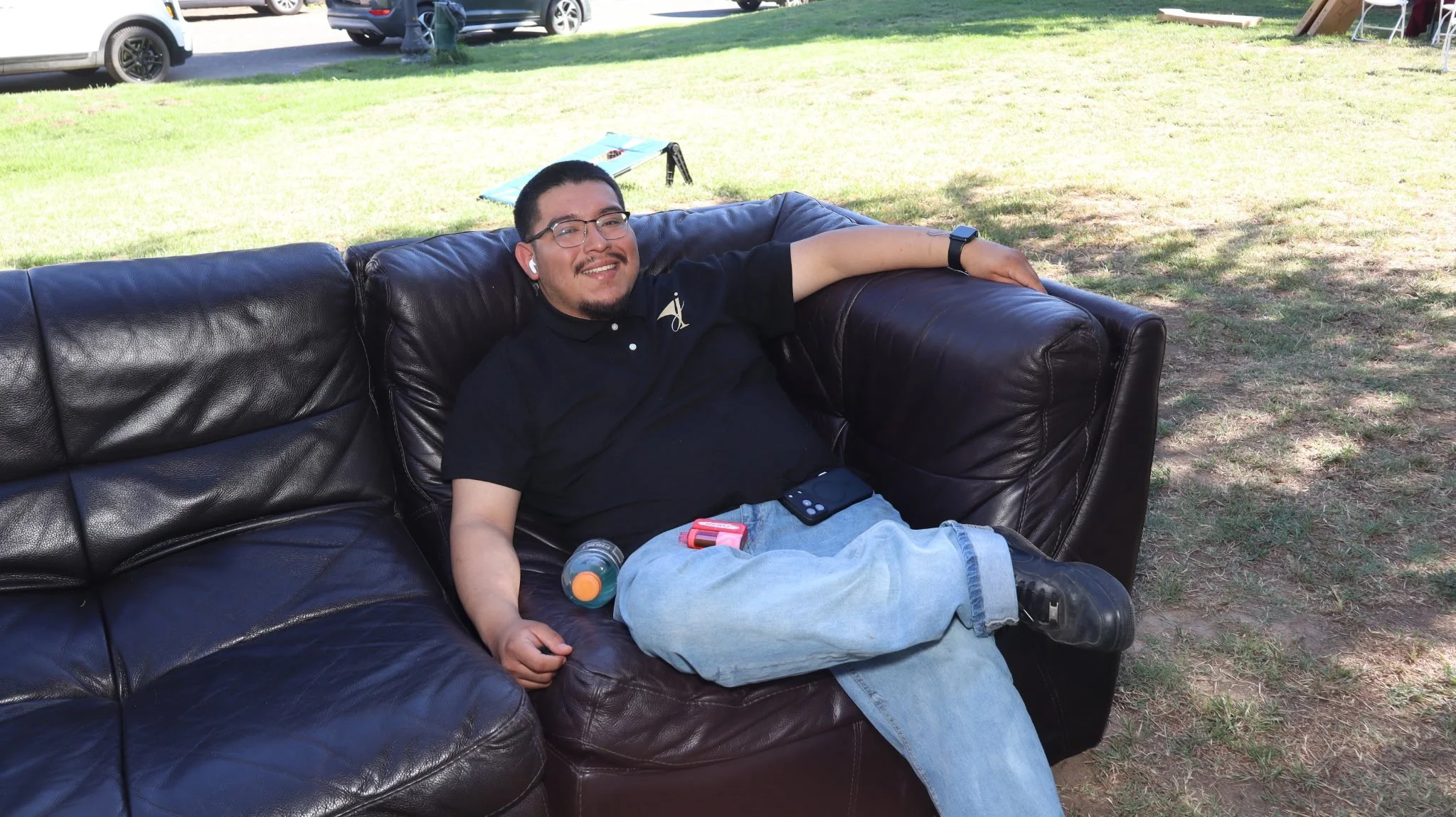 A young man with glasses, a black polo shirt, and jeans relaxing on a black leather couch outdoors on a grassy area in a park or yard, smiling at the camera with earbuds in his ears.