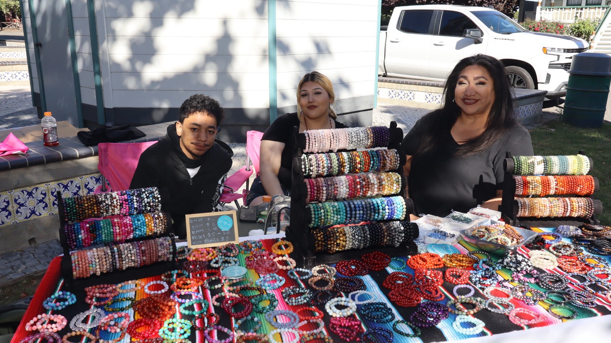 Three women sitting behind a display of colorful beaded jewelry at an outdoor market, with a white truck and a small white building in the background.