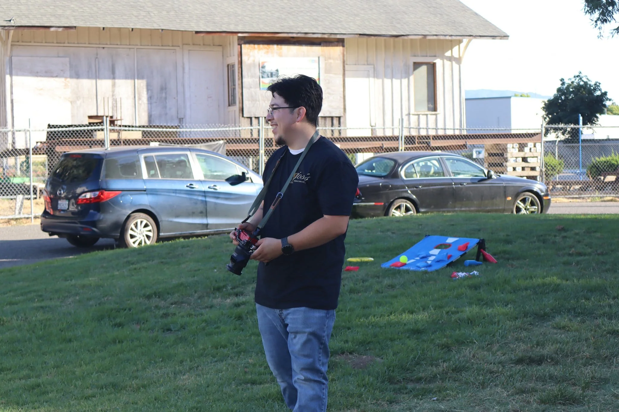 A man with glasses and dark hair holding a remote control standing on a grassy area near parked cars and a house in the background. There are toys and a game on the grass nearby.