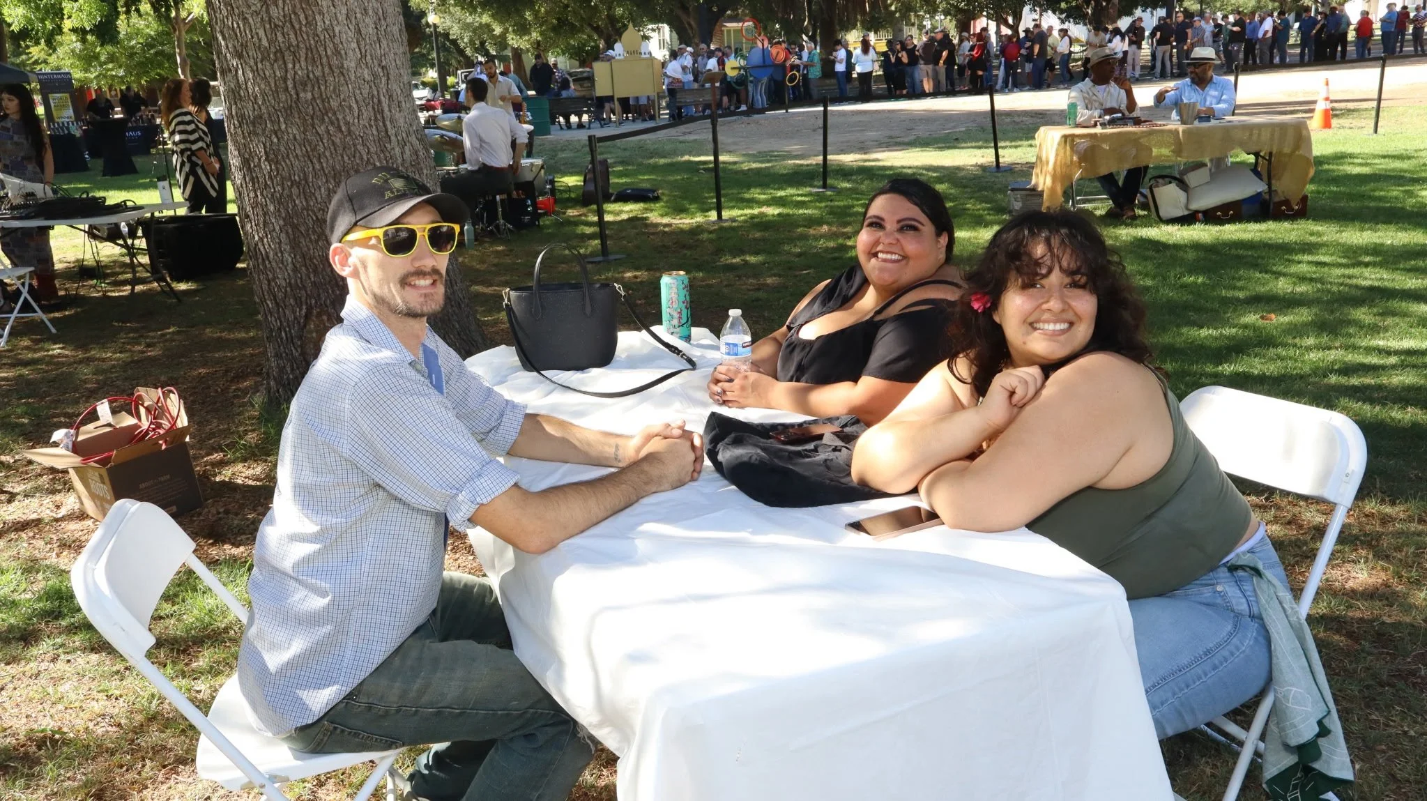 Three friends sitting at a table outdoors at a park, smiling, with a large tree in the background and a crowd of people in the distance.
