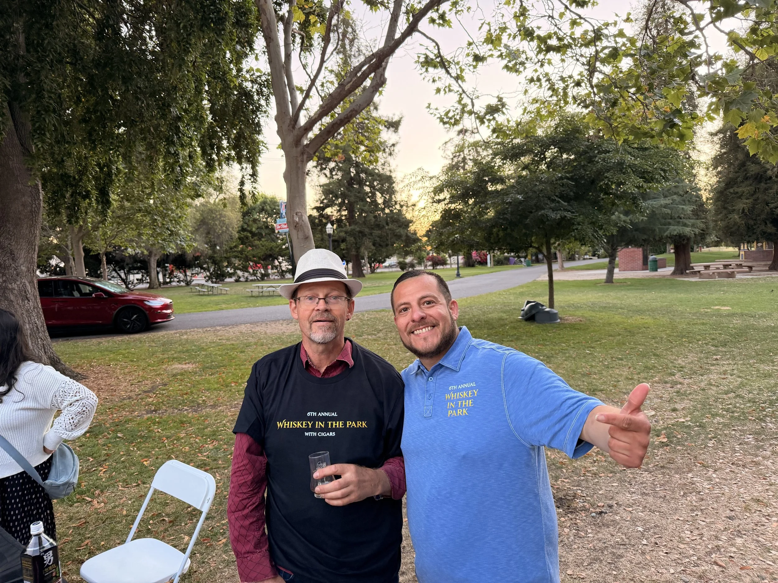 Two men smiling at an outdoor event in a park, one wearing a white hat and glasses, the other in a blue shirt, with trees and park benches in the background.