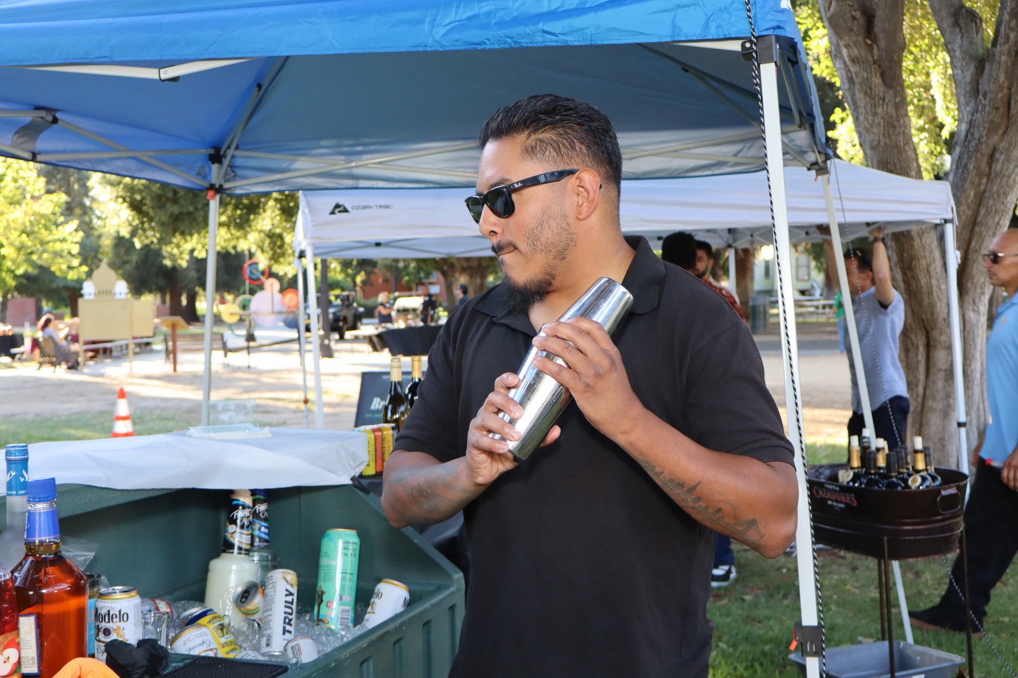 A man with dark hair, beard, and sunglasses holding a cocktail shaker at an outdoor social event with tents, trees, and people in the background.