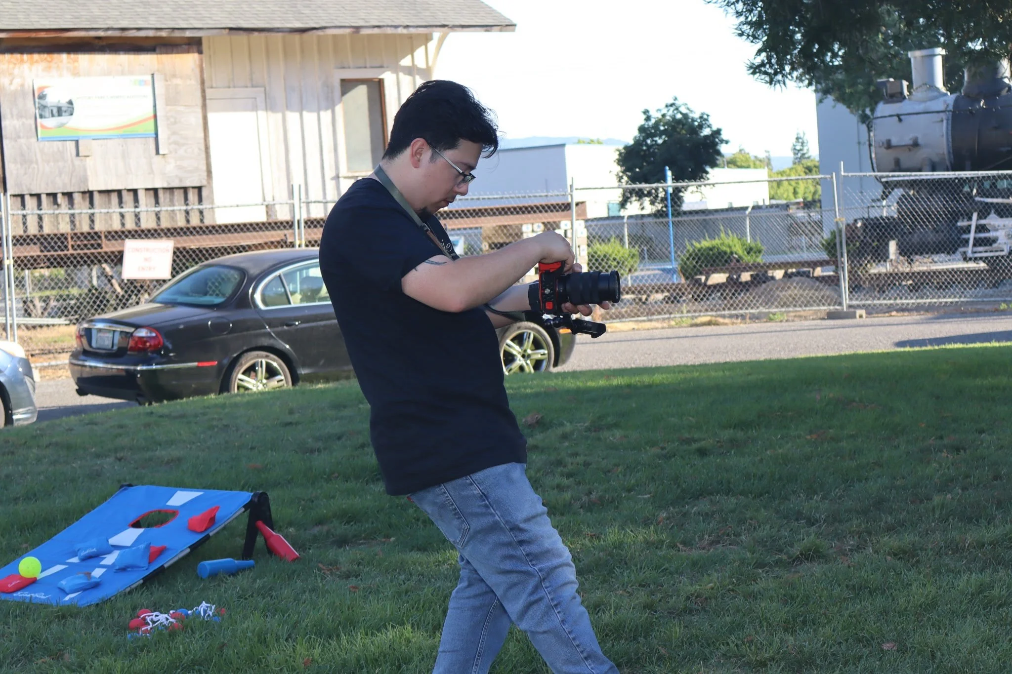 A man is standing outdoors on the grass, setting up a tennis training aid with a blue board and colorful balls, near parked cars and a wooden building.