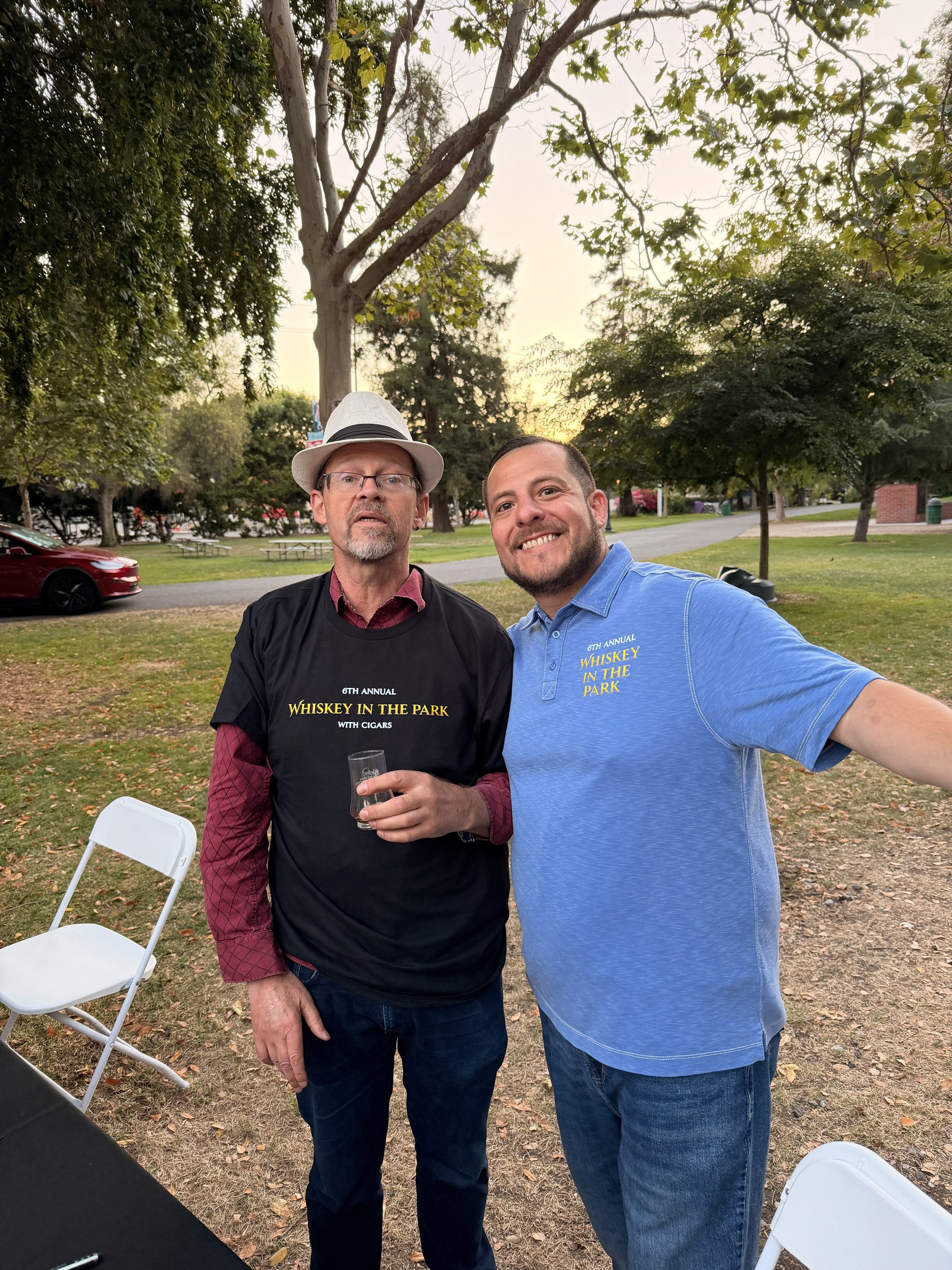 Two men take a selfie outdoors in a park during sunset. The man on the left is wearing glasses, a fedora, a black t-shirt with yellow text, and is holding a drink. The man on the right has short hair, is smiling, and is wearing a blue polo shirt with