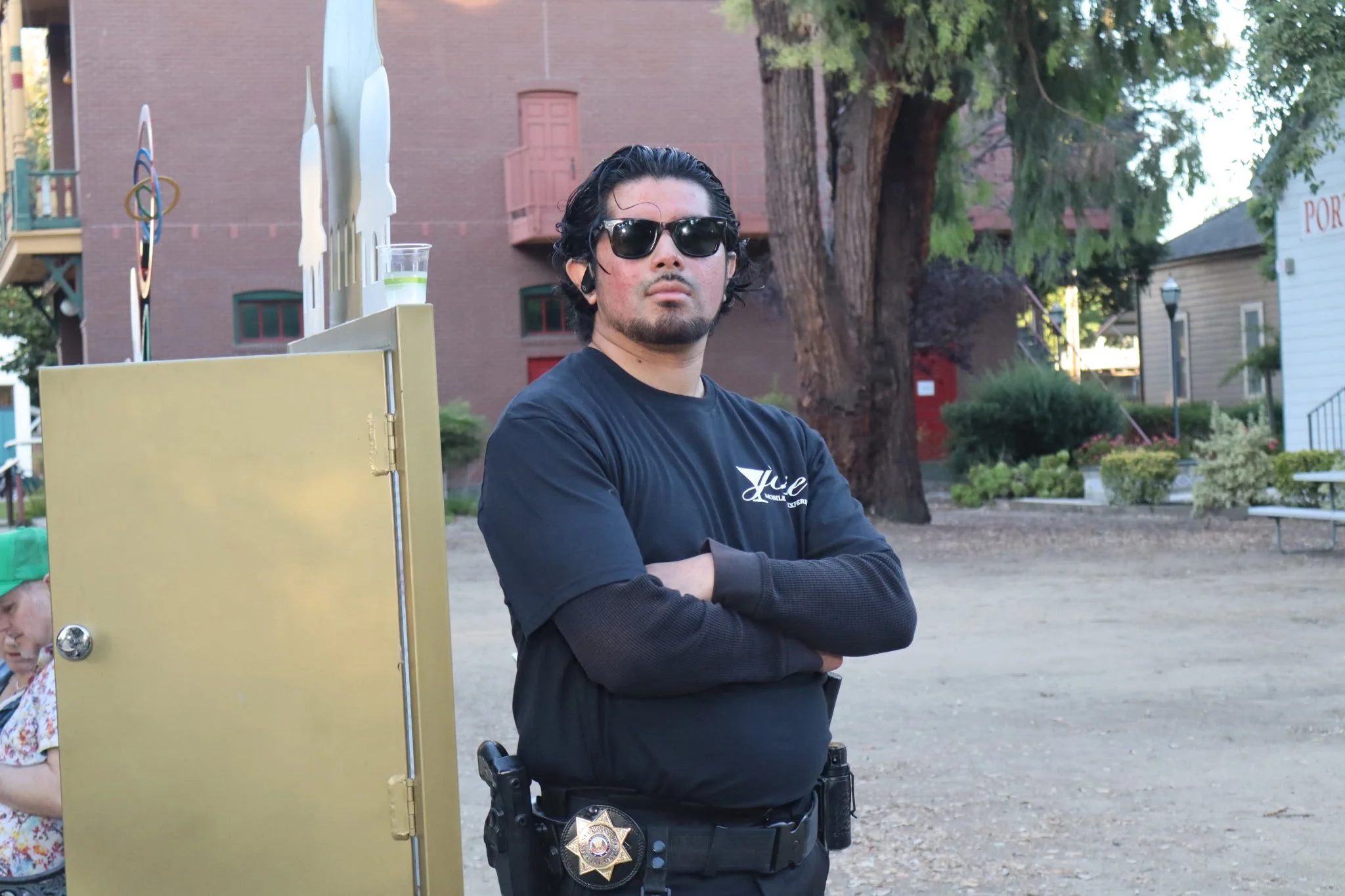 A man with dark hair, sunglasses, and a goatee, wearing a black shirt and black long-sleeve shirt underneath, stands with his arms crossed in an outdoor park. Behind him is a large tree, residential buildings, and sports equipment, including a basket