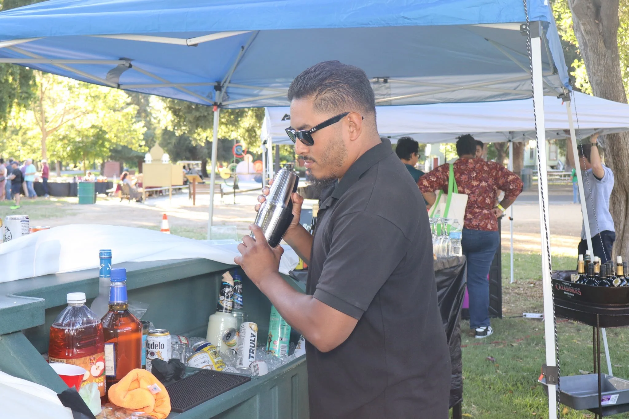 A man wearing sunglasses and a black shirt is behind a table with various bottles and cans, likely at an outdoor event or market. He is holding a shaker and standing under a blue canopy with other people and trees visible in the background.