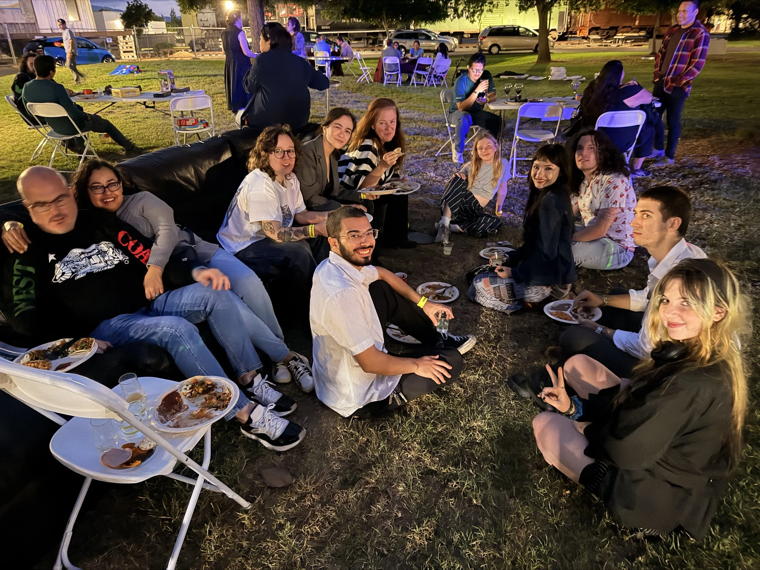 A group of diverse people gathered outdoors at night for a social event, sitting on the grass and on chairs, eating food, with tables and additional groups of people in the background, illuminated by artificial lighting.