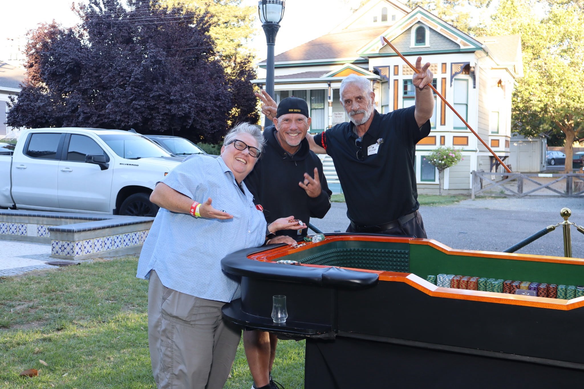 Three people smiling at a yard sale or outdoor event, standing near a grand piano filled with colored poker chips. One person is holding a glass, another is making a peace sign, and the third is holding a coin. In the background, there is a white veh
