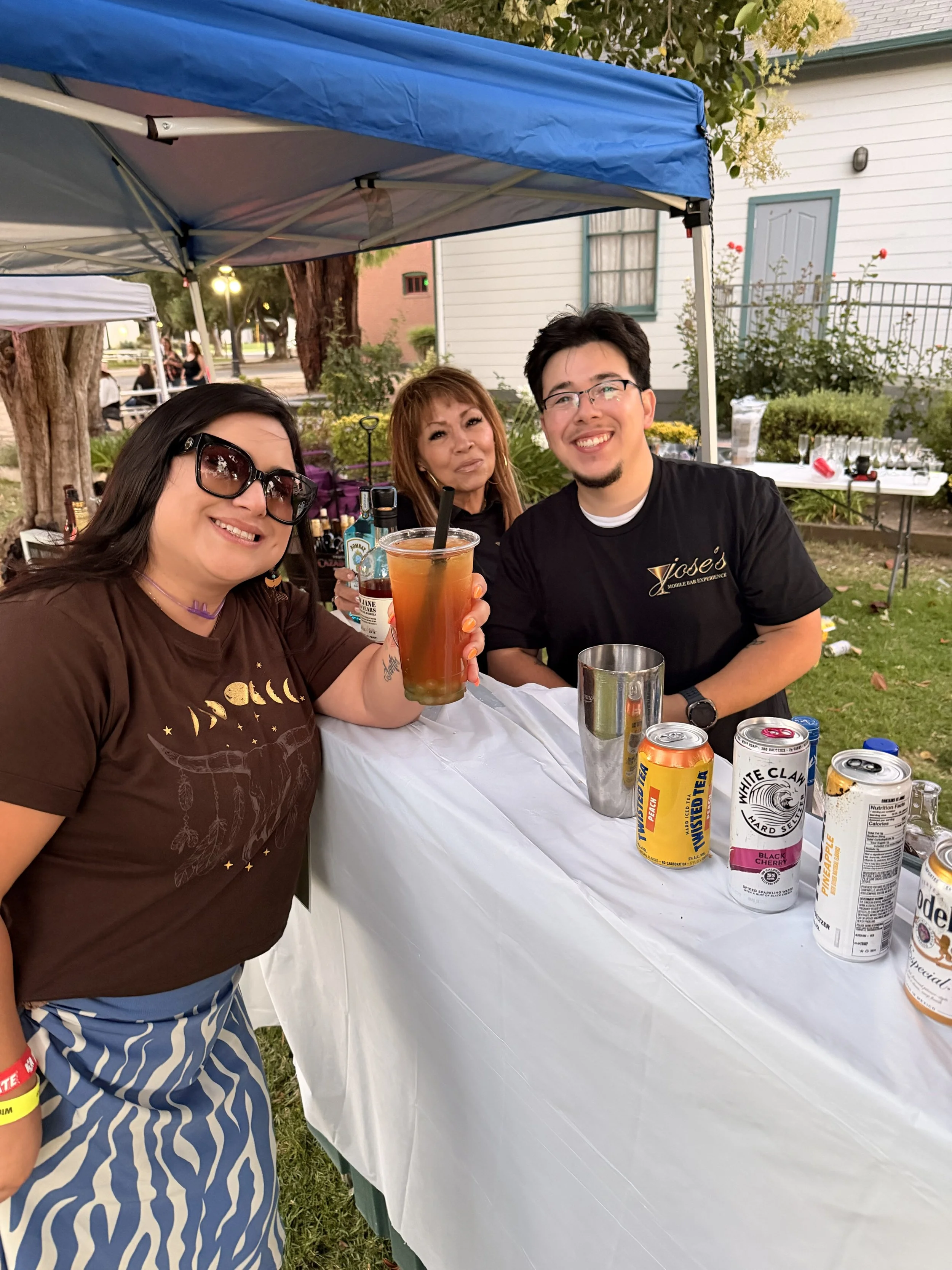 Three people sitting at a food or drink stand outdoors, smiling and holding drinks, with bottles and cans on the table.