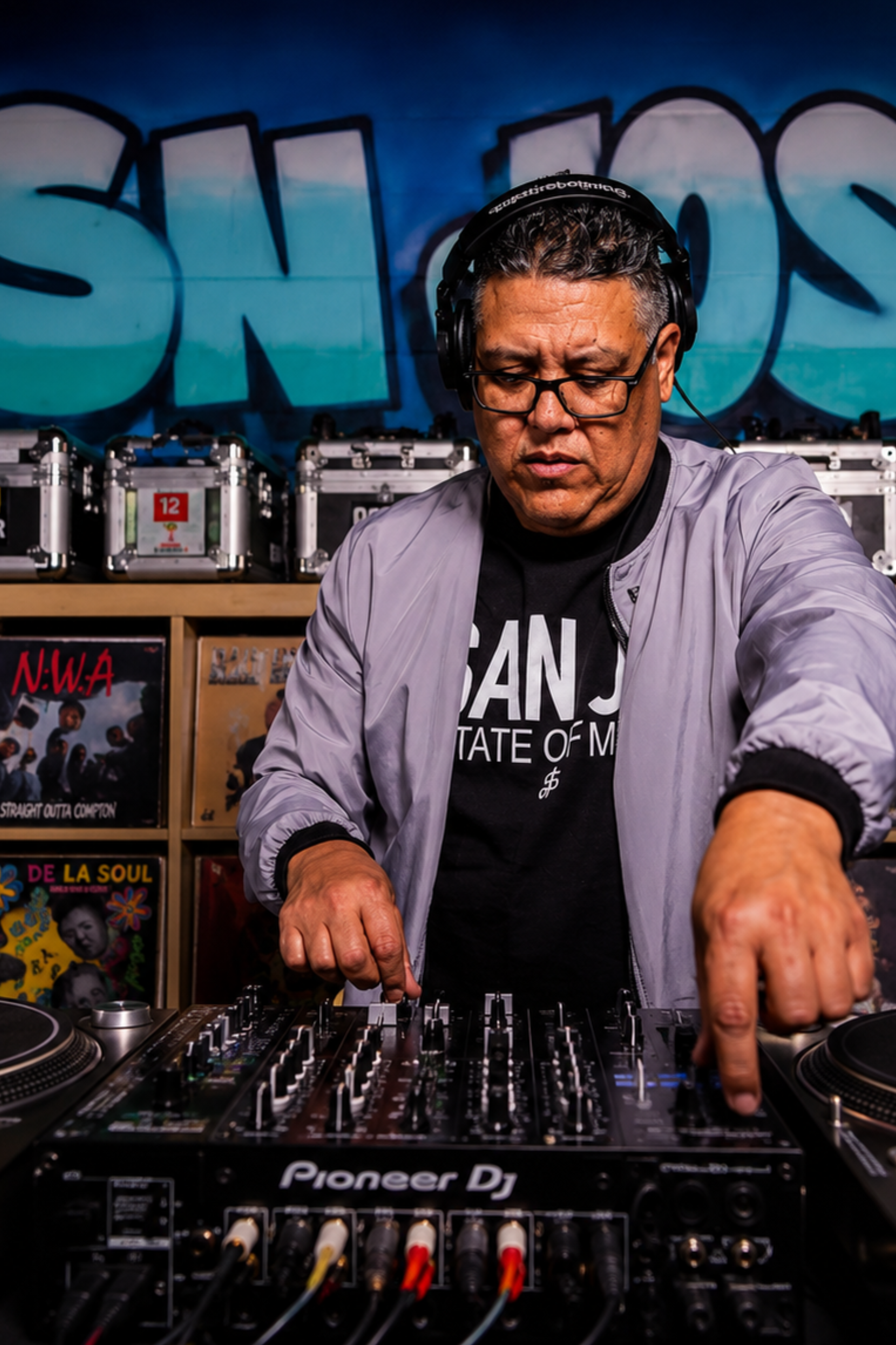 A man wearing glasses and headphones DJs with a Pioneer DJ mixer in a room decorated with graffiti and records.