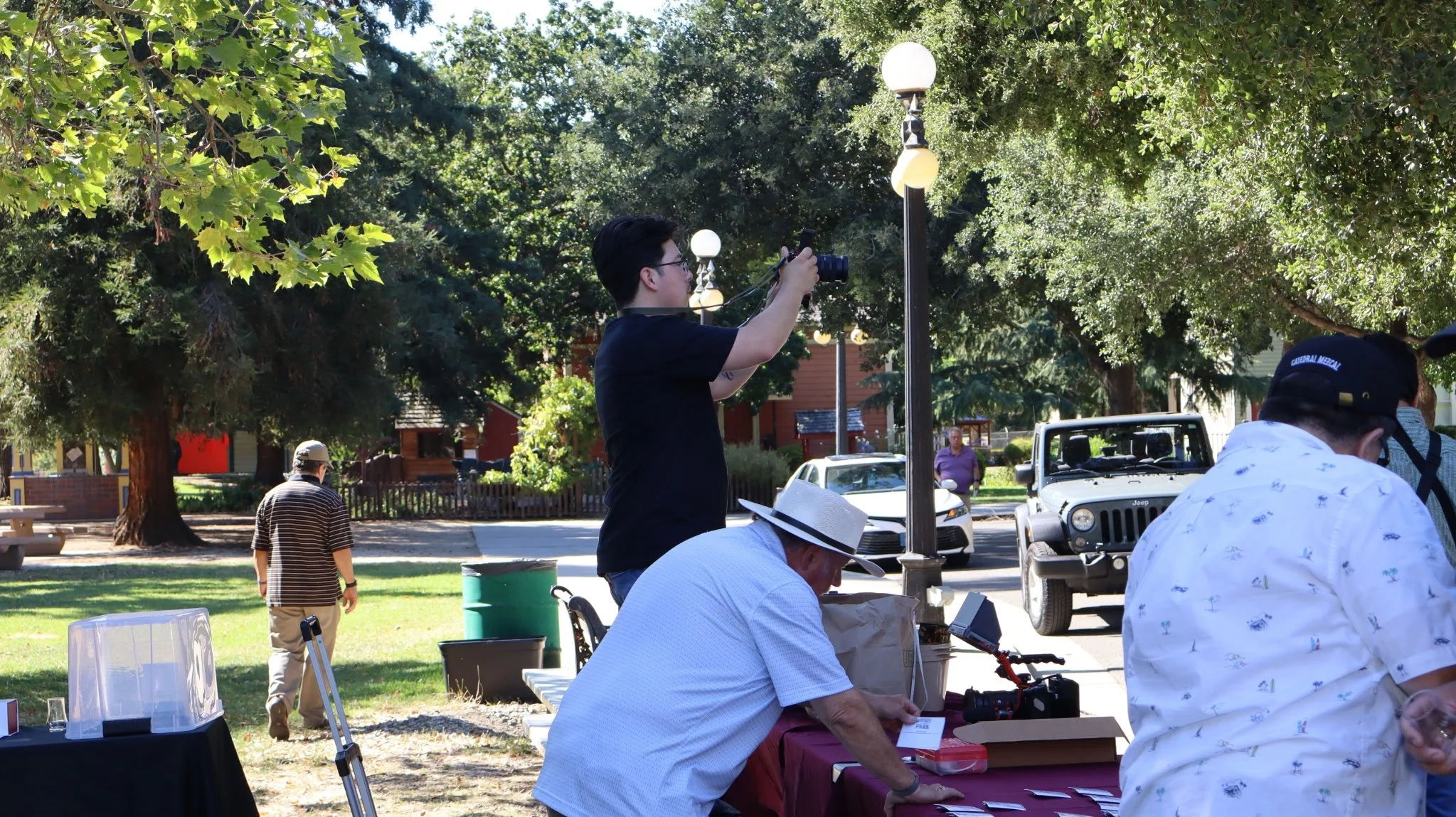 A young man taking a photo with a camera in a park during daytime. People are sitting and walking around with trees, streetlamps, and parked cars in the background.