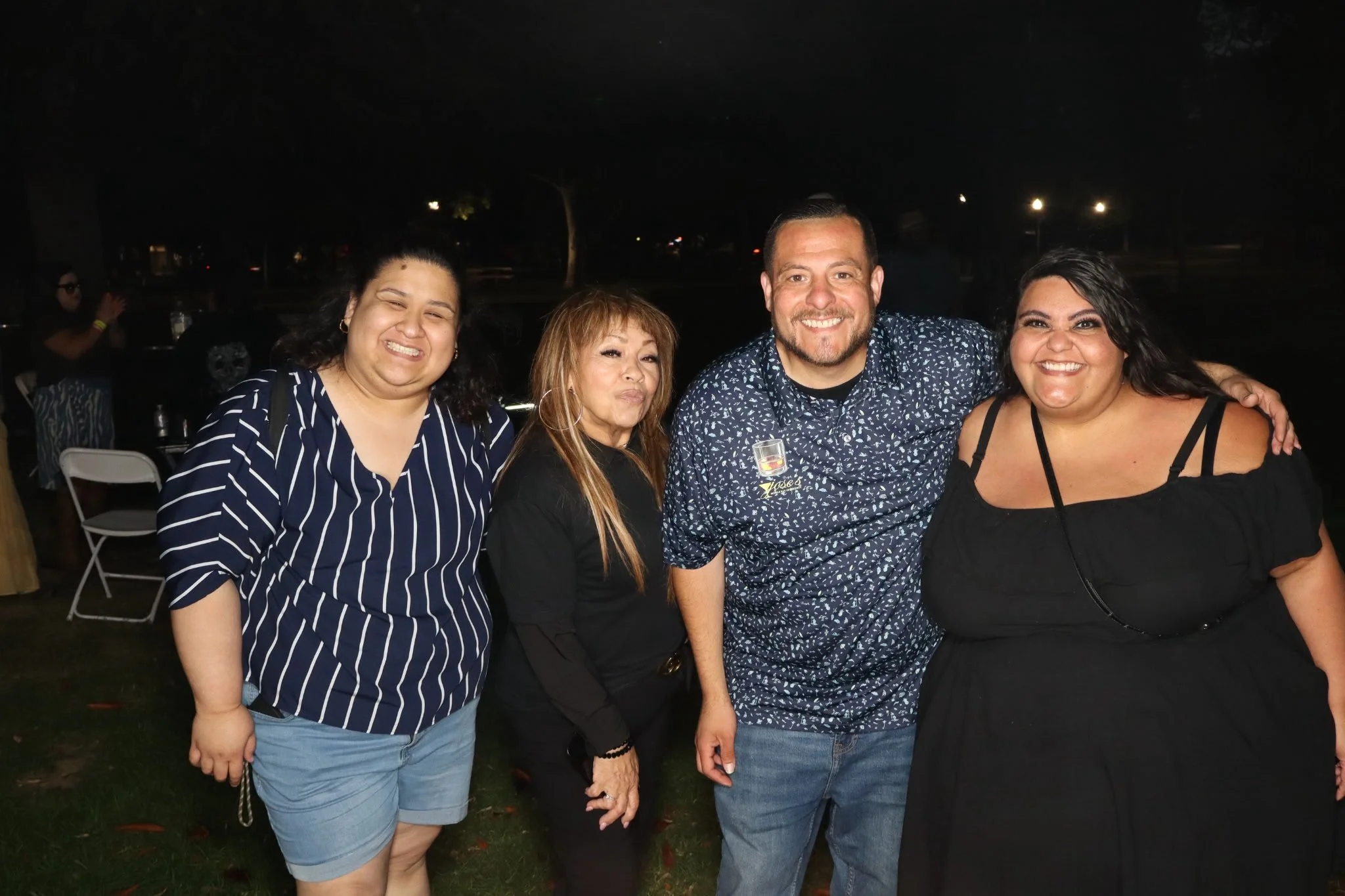 Four people smiling and posing for a photo at an outdoor event during nighttime, with dark background and some lights in the distance.