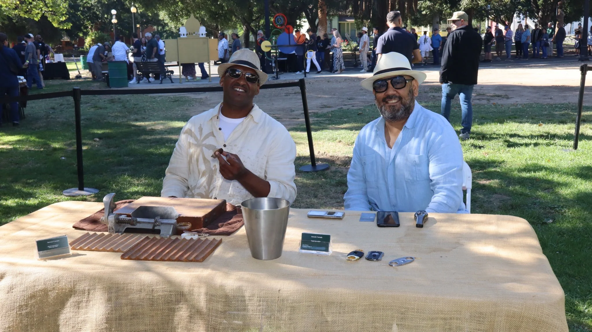 Two men wearing hats and sunglasses sitting at a table outdoors during a daytime event with a crowd in the background. The table has various objects, including a metal cup, velvet chamois, and electronic devices.