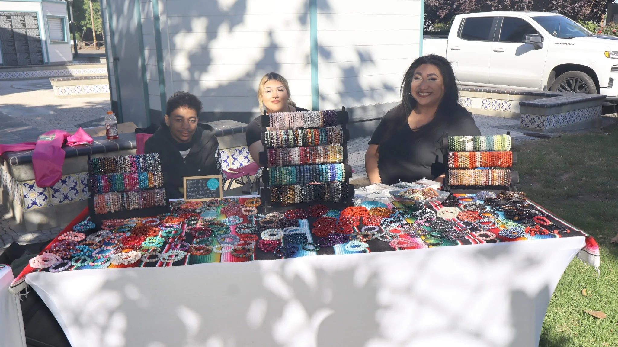 Three women sitting behind a table displaying colorful jewelry, including bracelets and necklaces, at an outdoor market stall with a white truck in the background.