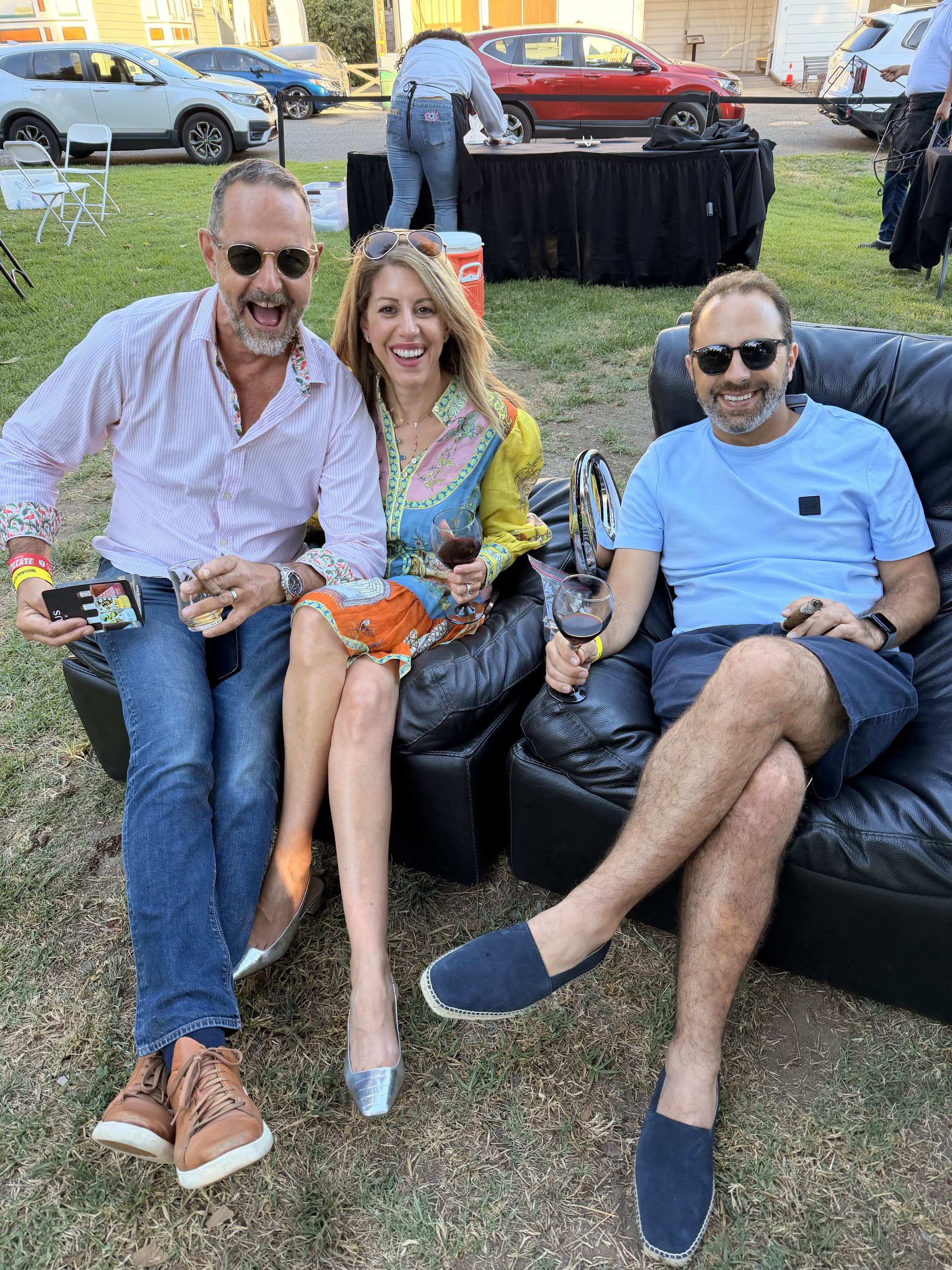 Three adults sitting on a black leather couch outdoors at a social gathering, smiling at the camera, each holding a glass of red wine. Behind them are parked cars and a person standing at a table.
