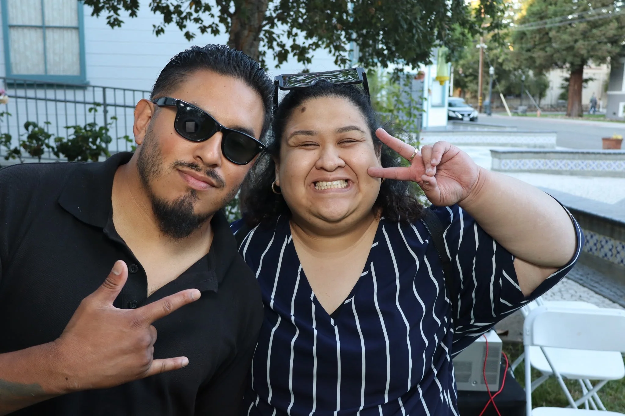 Two people posing outdoors, one man making a peace sign with his hand, both smiling and wearing sunglasses.