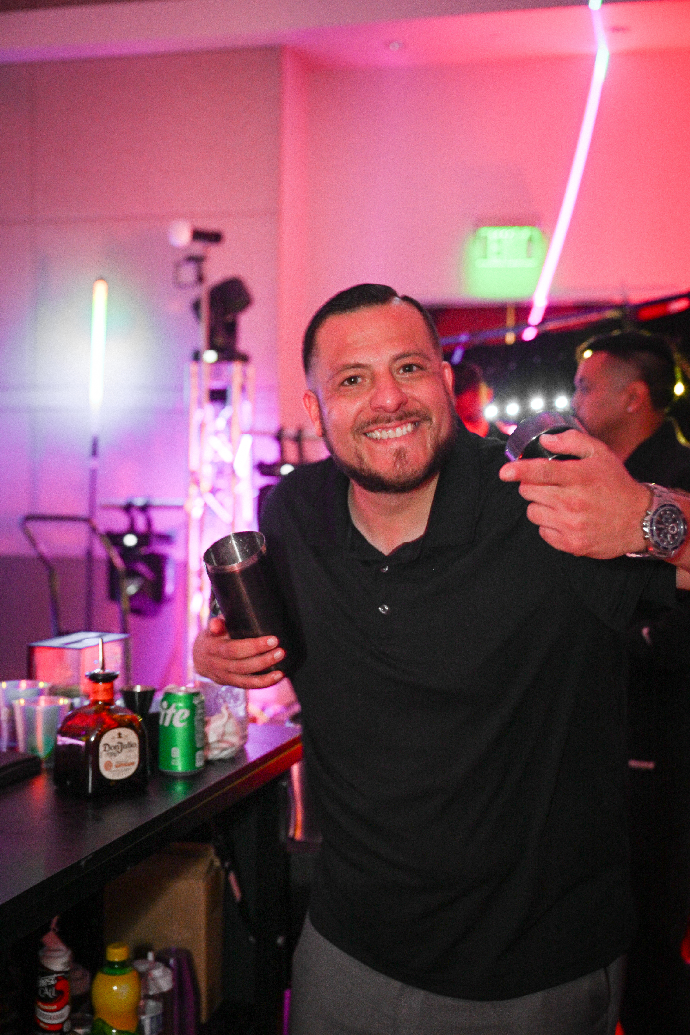 A smiling man holding a cocktail glass at a party, standing behind a bar with various bottles and cans, with colorful lighting and a blurred background of other people.