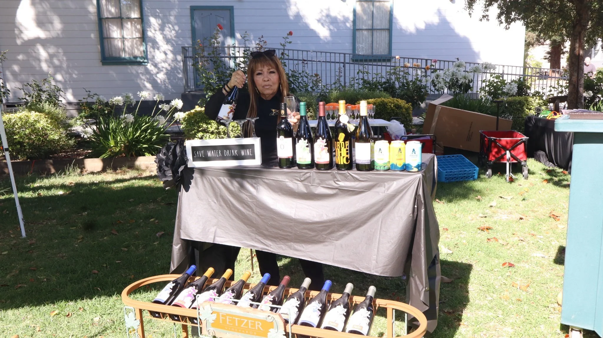 A woman standing behind a table selling wine outdoors. The table has several bottles of wine, a box of wine cartons, and a sign that says 'Save Water Drink Wine.' In front of the table, there are more wine bottles on a rack. The scene is set in a gar