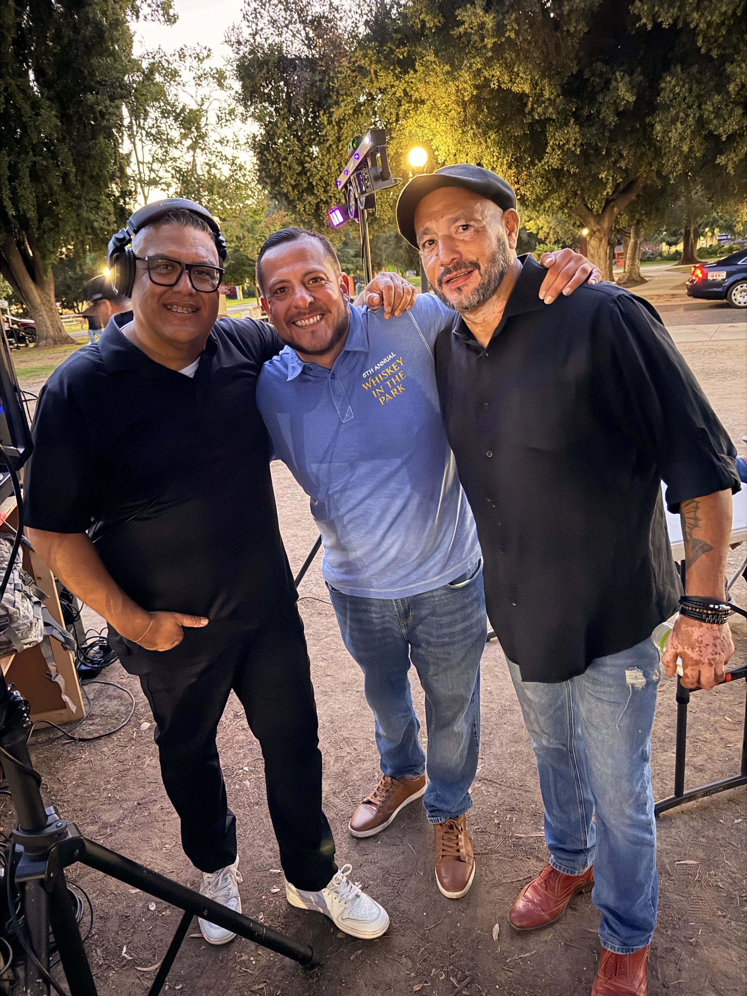 Three men standing closely together outdoors at sunset, smiling for a photo. One man on the left is wearing glasses, headphones, a black shirt, and white sneakers. The man in the middle is wearing a light blue shirt with yellow text, jeans, and brown