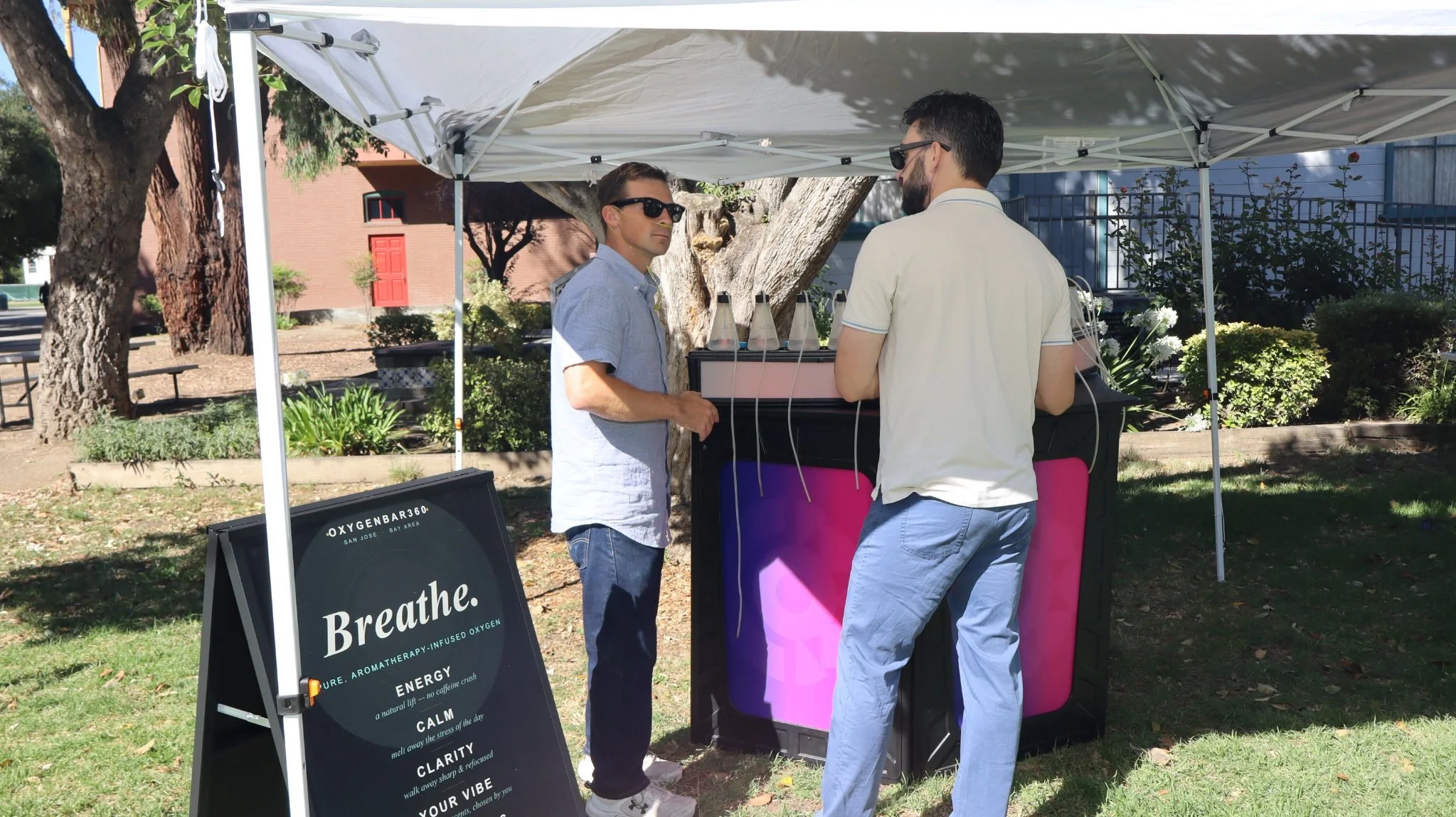 Two men wearing sunglasses standing under a white canopy at an outdoor event, with a sign promoting breathing exercises and oxygen therapy nearby, surrounded by trees and bushes.