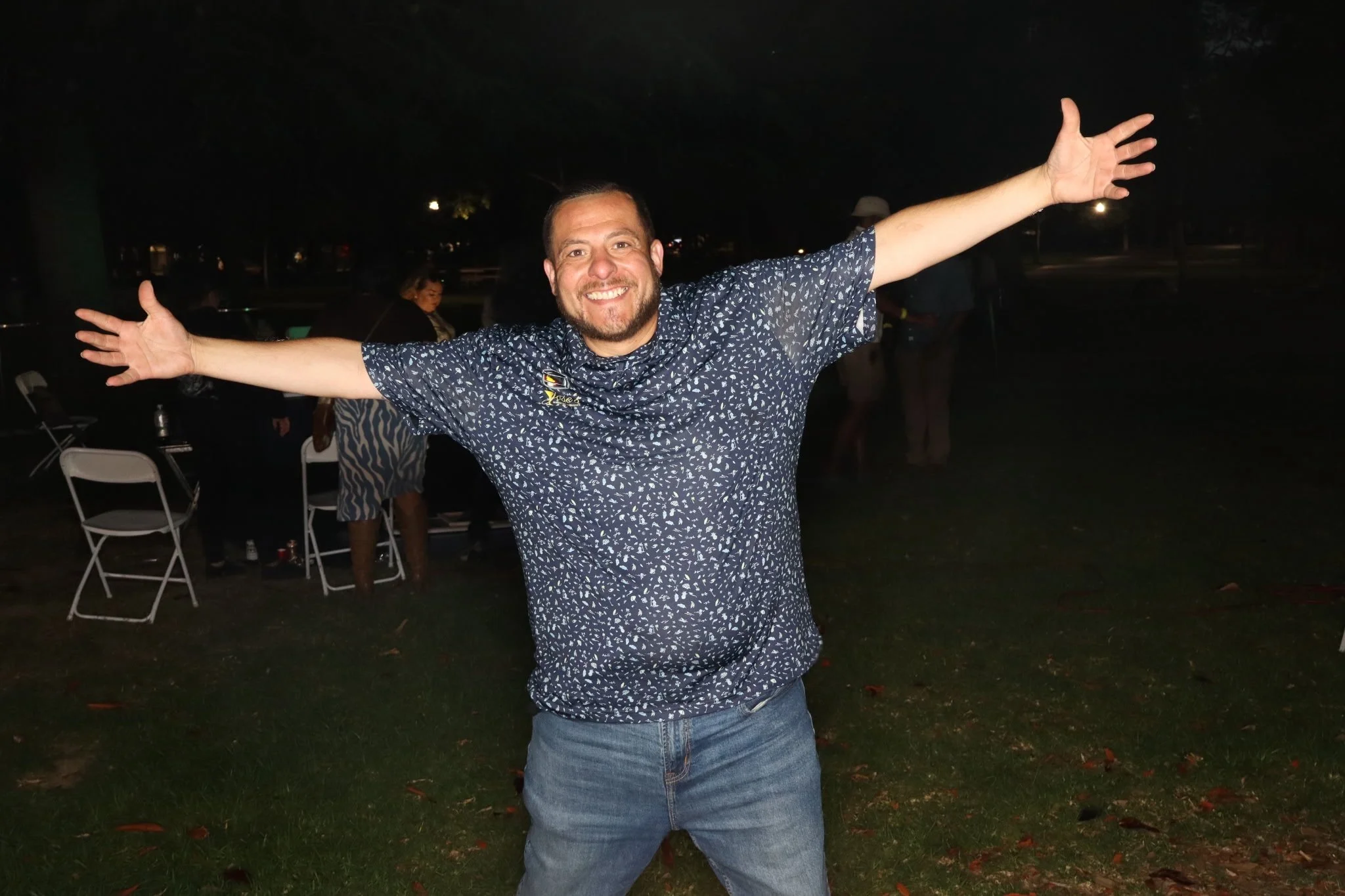 A man smiling with arms outstretched outdoors at night, wearing a patterned shirt and jeans.