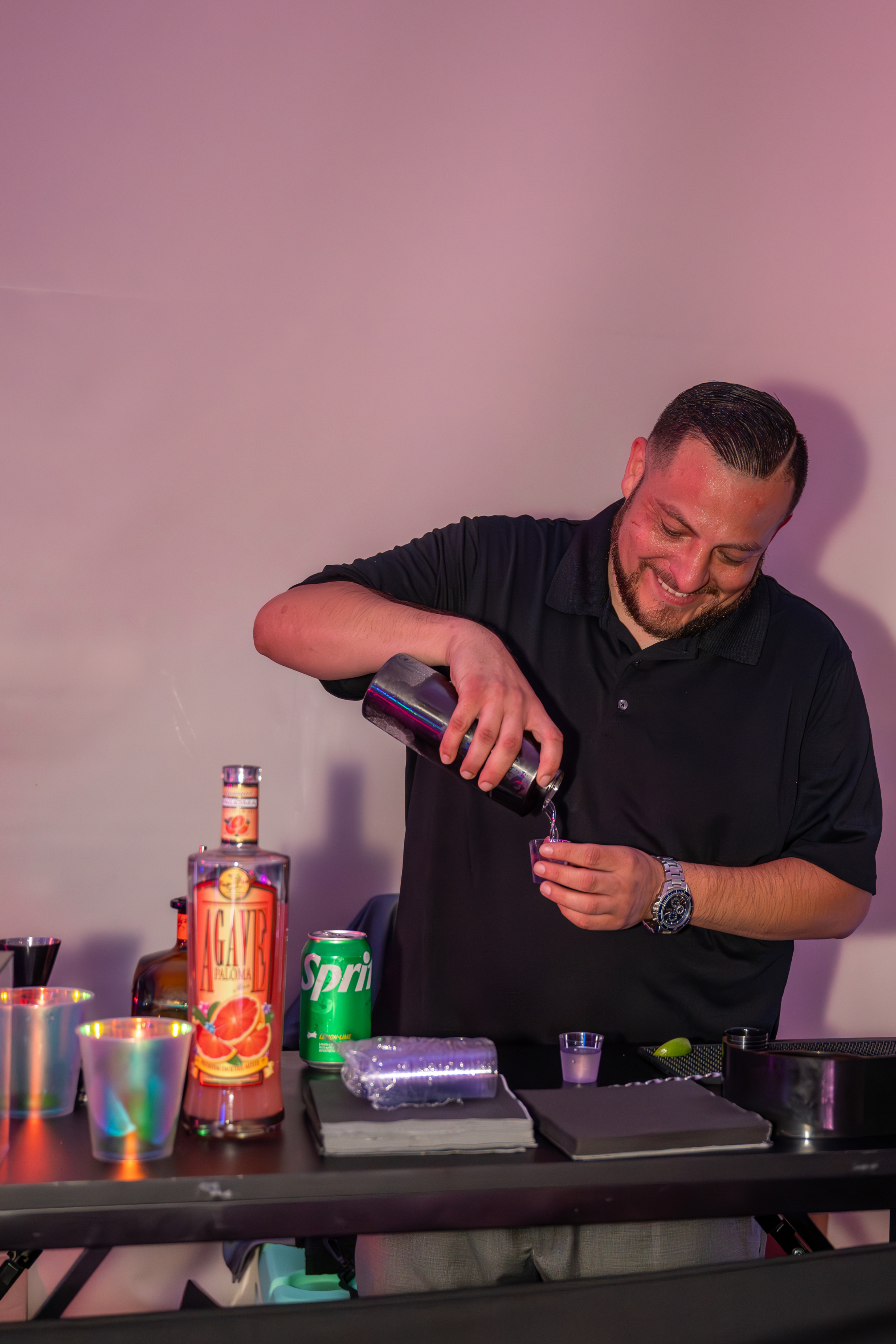 A man with dark hair and a beard, smiling, pouring a clear liquid into a small glass, standing behind a bar or drink preparation area with various bottles, cans, and bar tools on the counter.